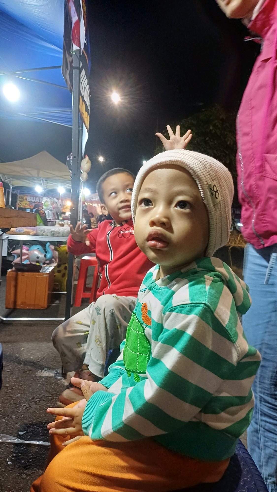 A low-angle, medium shot captures two young children sitting outdoors at night, likely at a night market in Kowangan, Indonesia. The child in the foreground, wearing a white beanie and a striped green and white hoodie with a green turtle appliqué, is looking upwards with a neutral expression. Their hands are clasped on their lap, which is covered by an orange fabric. Behind them, another child in a red zippered jacket and patterned pants looks towards the left, with their right hand raised slightly. The background is dimly lit, with hints of market stalls, lights, and trees. A blue canopy is visible overhead. The overall scene suggests a casual, nighttime outdoor gathering.