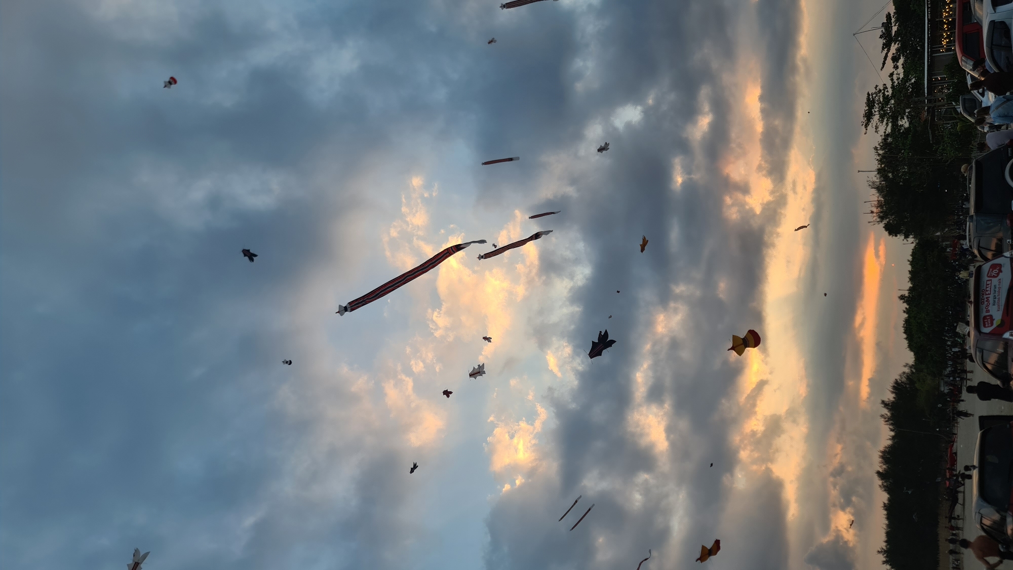 The image depicts a wide-angle view of a beach festival in Denpasar, Indonesia, with numerous kites of various shapes and sizes flying in a cloudy, dusky sky. The sky is a mix of dark gray clouds and patches of soft, warm light, suggesting either sunrise or sunset. The clouds are thick and textured, creating a dramatic backdrop for the kites.
The kites are the primary subjects, scattered across the frame. Some are long, striped banners, while others are shaped like birds or more abstract forms. Their silhouettes are visible against the brighter parts of the sky.
In the foreground, the scene transitions to a sandy beach area with a line of trees and vegetation separating it from a road. Several cars are parked on the side of the road, with some people gathered around them. A few individuals are visible on the beach, some walking and others standing. The overall atmosphere appears to be festive and active. The lighting is dim, consistent with twilight hours, and highlights the warm tones in the sky.