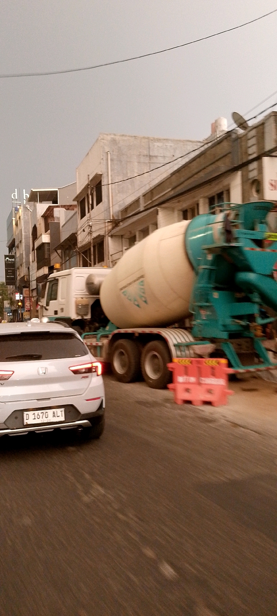 A white Honda car with license plate D 1670 ALT is captured in motion on a street in Bandung City, Indonesia. The car is followed by a cement mixer truck, also in motion, with a large beige drum and a teal-colored mixing mechanism. The truck is partially obscured by the car in the foreground. In the background, there are several multi-story buildings lining the street. The sky is overcast, suggesting a cloudy day. The scene appears to be a typical urban street with ongoing construction or transportation activities.