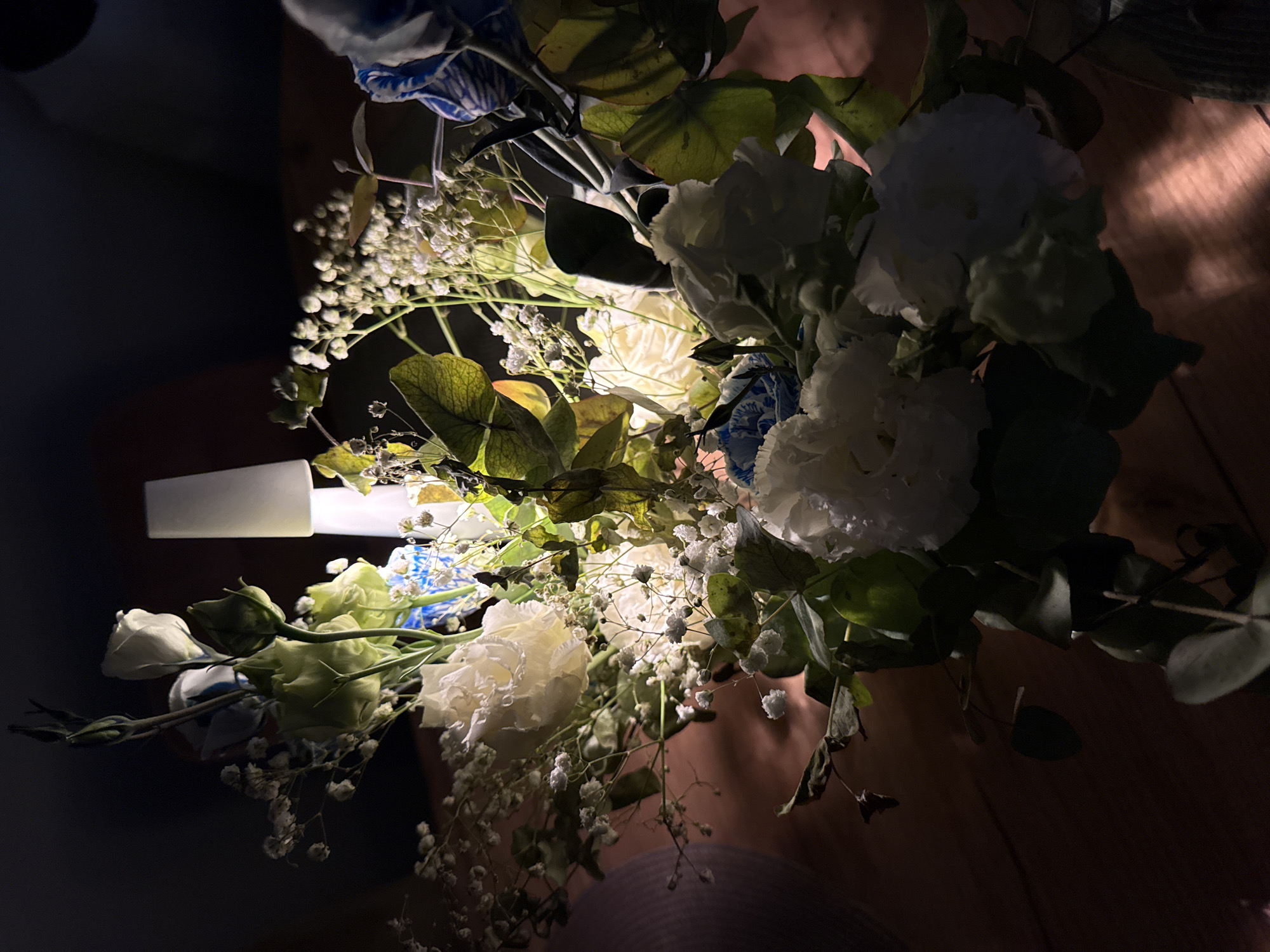 A close-up shot captures a bouquet of white and blue flowers, accented with delicate white baby's breath. The arrangement is illuminated by a soft, warm light, likely from a nearby lamp, casting gentle shadows and highlighting the textures of the petals and leaves. The flowers are predominantly white, with some featuring subtle blue accents, and are clustered together with lush green foliage. The background is dark and indistinct, suggesting an indoor setting. The bouquet is placed on a wooden surface, visible as a warm-toned, textured background. There is no evidence of people or any specific location in Sofia, Bulgaria.