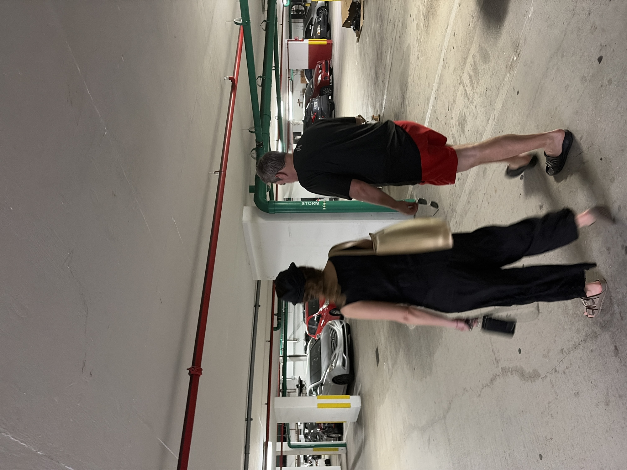 The image depicts two individuals walking in what appears to be a parking garage in Key Biscayne, United States of America. The first individual, closer to the camera, is wearing a black outfit and sandals, carrying a gold bag and a phone. The second individual, further away, is wearing a black shirt and red shorts, with black sandals. Various cars are parked in the background. The garage features overhead pipes, some painted red and green. The lighting is somewhat dim.