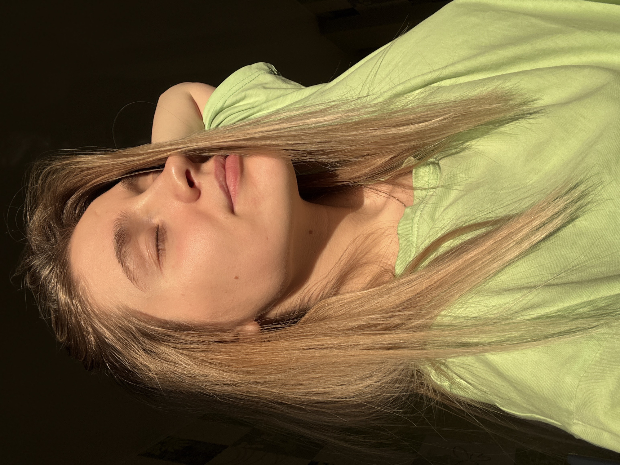 A young Caucasian female is depicted indoors, illuminated by a strong, warm, directional light source from the right. She has long, straight, light blonde or light brown hair that falls over her shoulders. Her head is tilted slightly upward and to the left, with her eyes closed and a relaxed facial expression. Her right arm is bent with the hand positioned behind her head, and the elbow is visible near her ear. She wears a plain, light green long-sleeved garment and a thin metallic chain necklace. The background is predominantly dark and out of focus, showing only vague, indistinct shapes, including a faint vertical lighter rectangle in the upper left. No specific identifying features of Izhevsk, Russia, are visible in the scene; it appears to be a generic interior space.