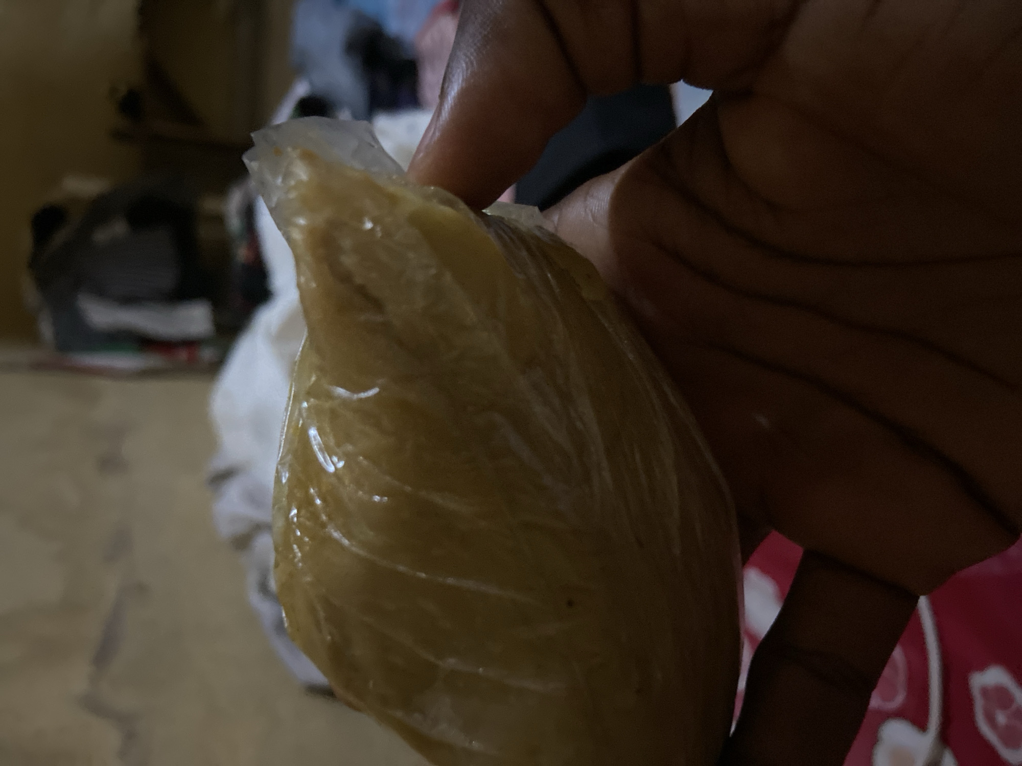 A person's hand is holding a food item wrapped in clear plastic. The food appears to be a solid, possibly a dough or paste, with a light brown or tan color. The plastic wrapping is slightly crinkled and reflects light. The background is blurred, but suggests an indoor environment with some clothing or fabric visible. The image does not contain any discernible landmarks or elements that would indicate a specific location within Jalingo, Nigeria.