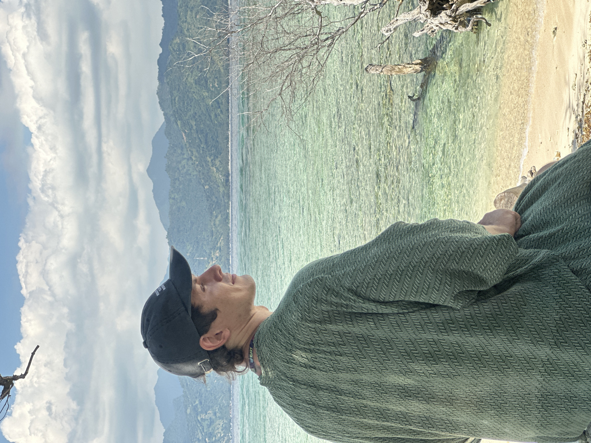 The image shows a person, likely a young adult male, wearing a black baseball cap and a patterned green shirt or sweater, looking upwards towards the sky. The scene appears to be set on a beach in an undetermined location in Indonesia. In the background, there's a calm, clear body of water with sandy shallows, and beyond that, a mountainous, forested landscape. A dead tree trunk is partially submerged in the water near the beach. The sky is overcast with clouds.