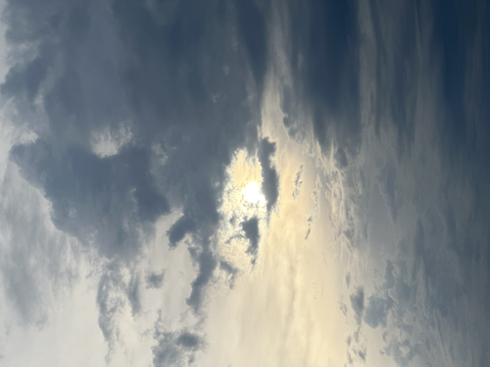 The image depicts a view of the sky with the sun partially obscured by clouds. The clouds are a mix of dark gray and lighter, brighter areas, suggesting a partly cloudy or overcast day. The location is Fethiye, Turkey, but there are no landmarks or other environmental elements visible in the image to confirm the city. There are no people, subjects, objects, actions, or interactions present within the scope of this image.