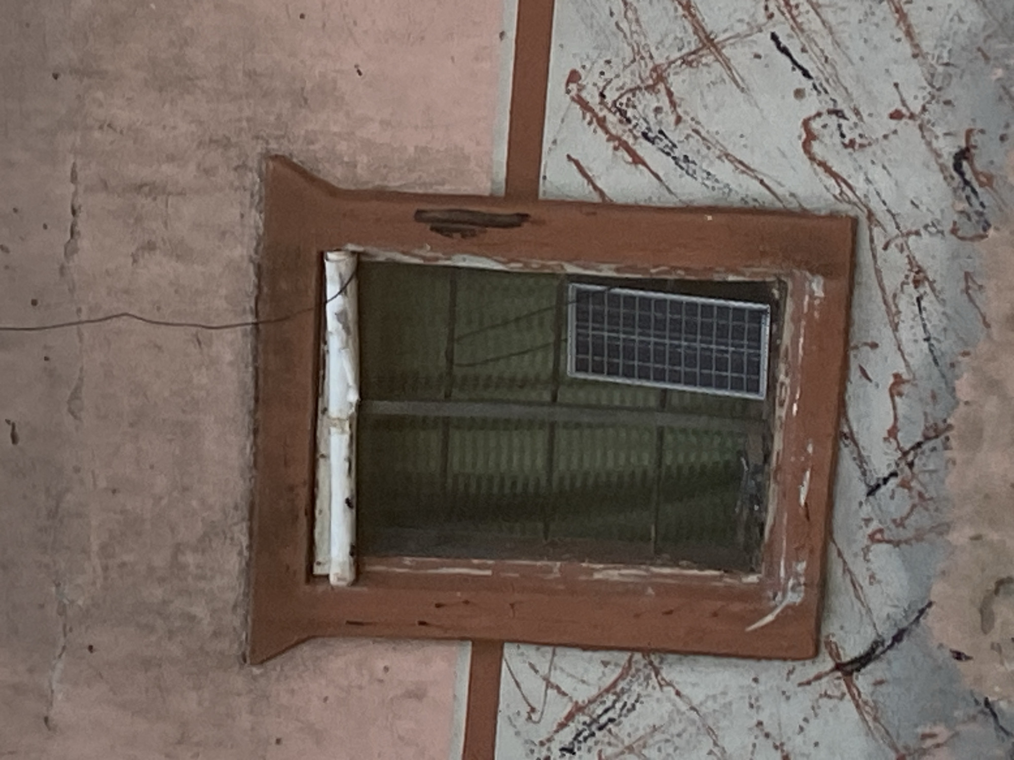 The image shows a close-up of a window with a small solar panel visible inside. The window frame is made of a reddish-brown material and appears weathered. Above the window, there is a white rolled-up object, possibly a shade or blind. The wall surrounding the window is painted in a pale pinkish-beige color and shows signs of age and wear, with some cracking and discoloration. The lower part of the wall has a decorative pattern of red and black streaks on a lighter background. A thin black wire runs down from the top of the image towards the window. The solar panel is rectangular with a grid of cells. The overall scene suggests an outdoor setting on a building. The image is too cropped to determine specific details of the surrounding environment or people.