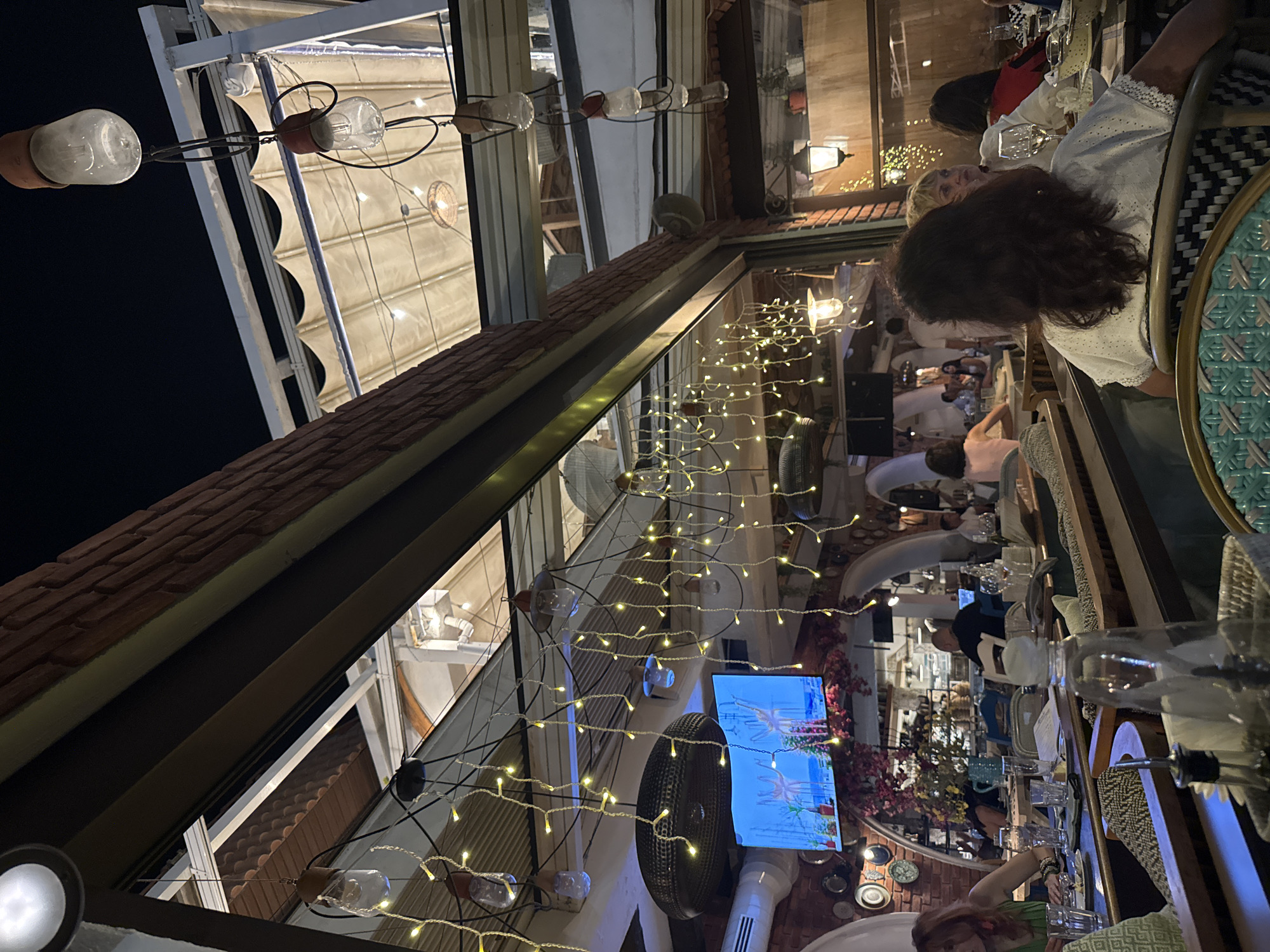 This is an outdoor dining area of a restaurant at night, likely located in Varna, Bulgaria. The scene is illuminated by string lights and decorative bulbs hanging from the ceiling and from exposed beams. People are seated at tables, dining and conversing. A television screen is visible on a brick wall, displaying an image. The architecture suggests a covered patio or balcony, with exposed brick and a retractable awning overhead. Various dining settings, including plates, glasses, and bottles, are present on the tables. The overall atmosphere is one of a relaxed evening meal.