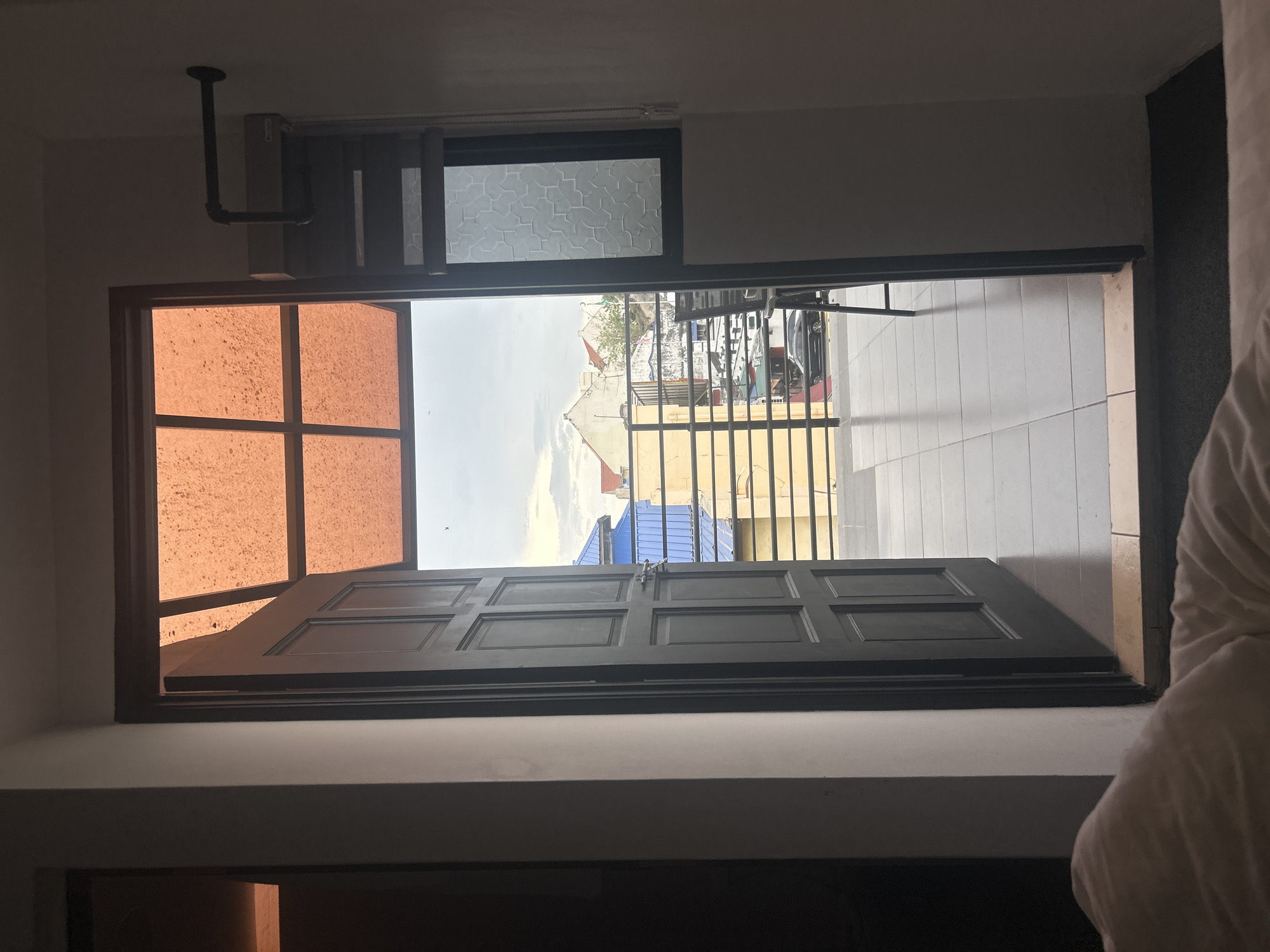 The image shows a view from inside a room, looking out through an open door and an awning to a street scene in George Town, Malaysia. The door is dark grey, paneled, and ajar. Above the door is an orange, translucent awning with a grid pattern. Beyond the door, a tiled balcony or walkway is visible, leading to a railing. Through the railing, buildings with blue and yellow roofs, and a white building with red accents are seen. A black car is parked on the street below. To the right of the door, a window with a textured, opaque glass pattern is visible. A metal pipe extends from the wall to the right of the awning. In the foreground, the edge of a white duvet or comforter is visible, suggesting the photo was taken from a bed. The sky is overcast.