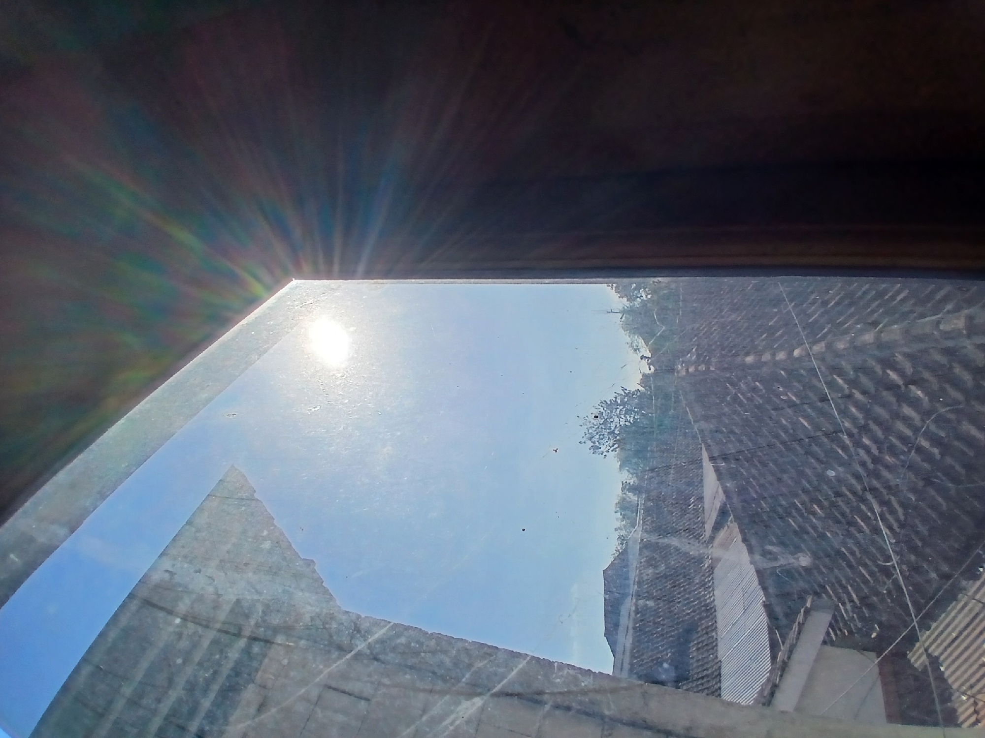 The image is a sunlit view through a window overlooking a residential area, possibly in Tenggarang, Indonesia. The foreground features a dirty window pane with visible smudges and scratches, dividing the scene. The left side of the window shows a bright sun with lens flares, casting rays into the frame. Below the sun, a blue sky is visible with some distant trees and scattered dark specks, likely dust on the window. The right side of the frame is dominated by a dark, shadowed surface, possibly the interior of the room. The view outside the window reveals a dense arrangement of tiled rooftops and some corrugated metal roofs, characteristic of urban or suburban Indonesian architecture. The overall impression is that of a sunny day, with the light significantly filtered and distorted by the dirty window.