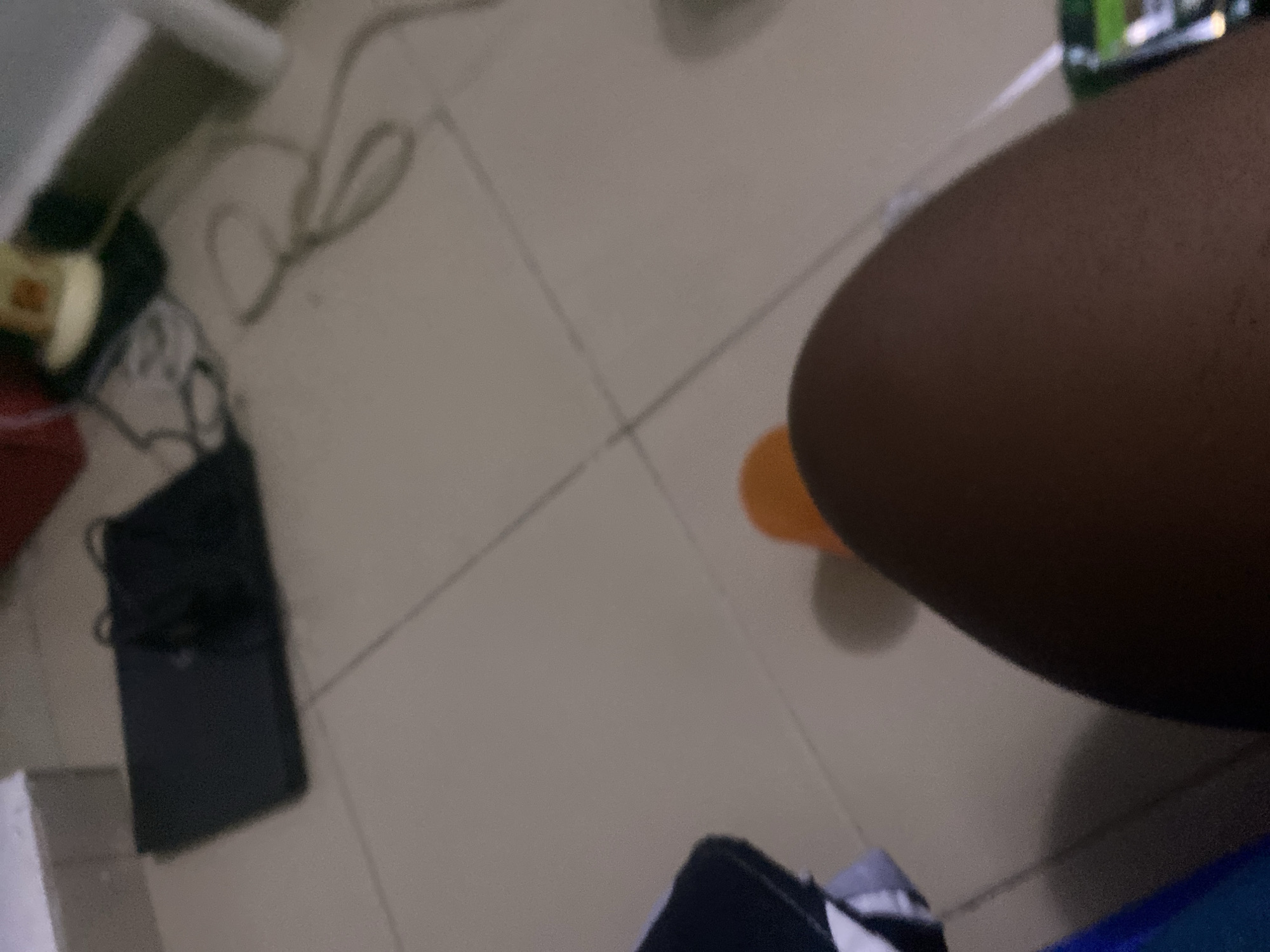 In a dimly lit room, a person's bare leg and thigh are prominent in the foreground, with skin appearing tanned. An orange slipper is visible on the floor near the leg. The background shows a tiled floor with faint grout lines. On the floor, a black electronic device, possibly a game console or laptop, is partially obscured by shadows, with cables trailing from it. Other indistinct objects and furniture are visible in the blurred background, suggesting a domestic or indoor setting. The image does not provide enough visual information to determine the specific location within the city of Howayi, Nigeria.