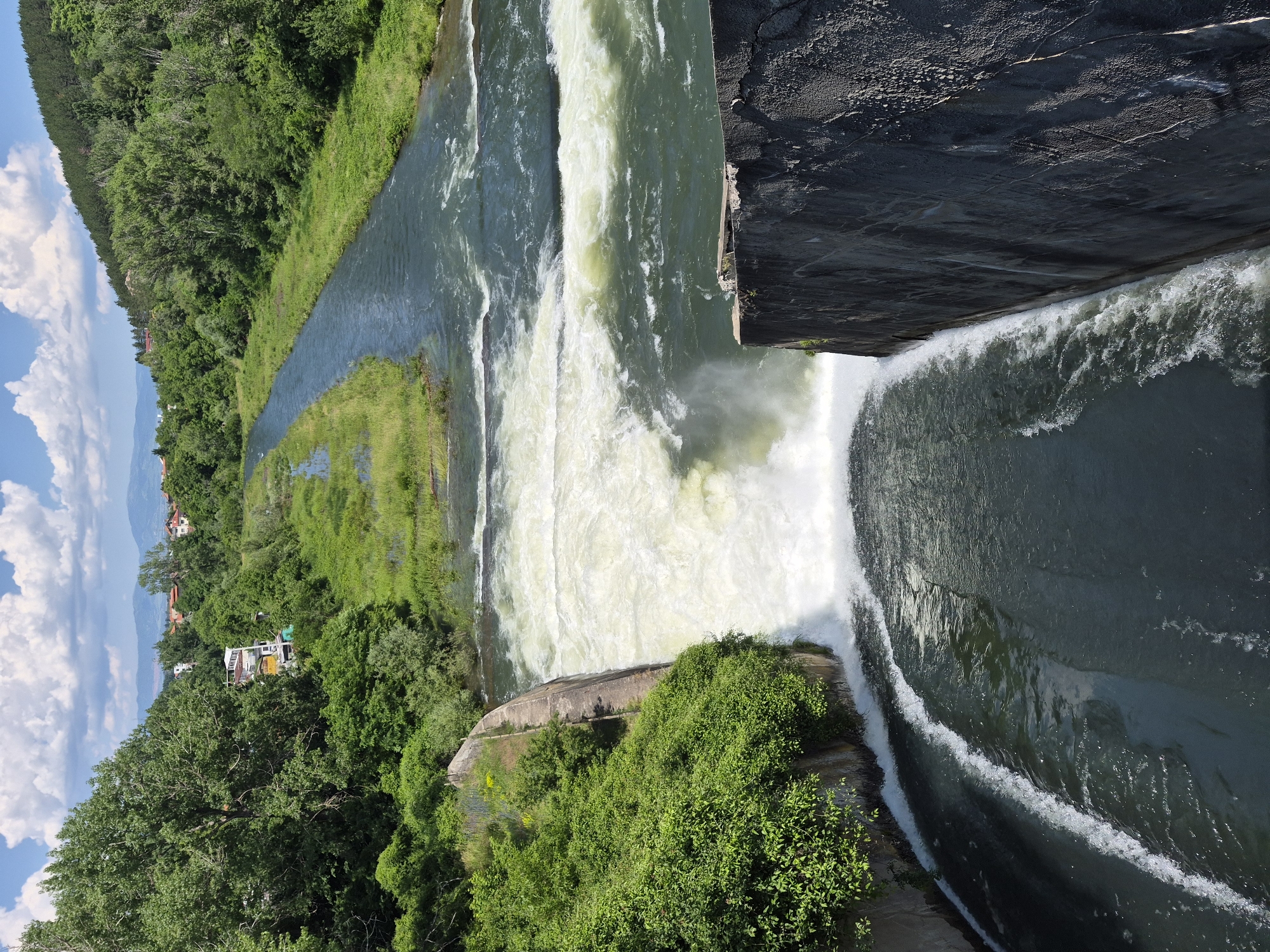 An overhead, medium-shot captures a raging river flowing over a dam with lush green vegetation on either side. The dam, constructed from rough concrete, is in the foreground, with white water cascading down its face and pooling below. The river, a murky green, is turbulent and wide, with smaller cascades and rapids visible upstream. The banks of the river are densely covered with various shades of green trees and plants, with some buildings and a distant mountain range visible in the background. The sky is a vibrant blue, dotted with fluffy white clouds. The scene appears to be in a natural or semi-natural setting, possibly a park or a rural area on the outskirts of a city.