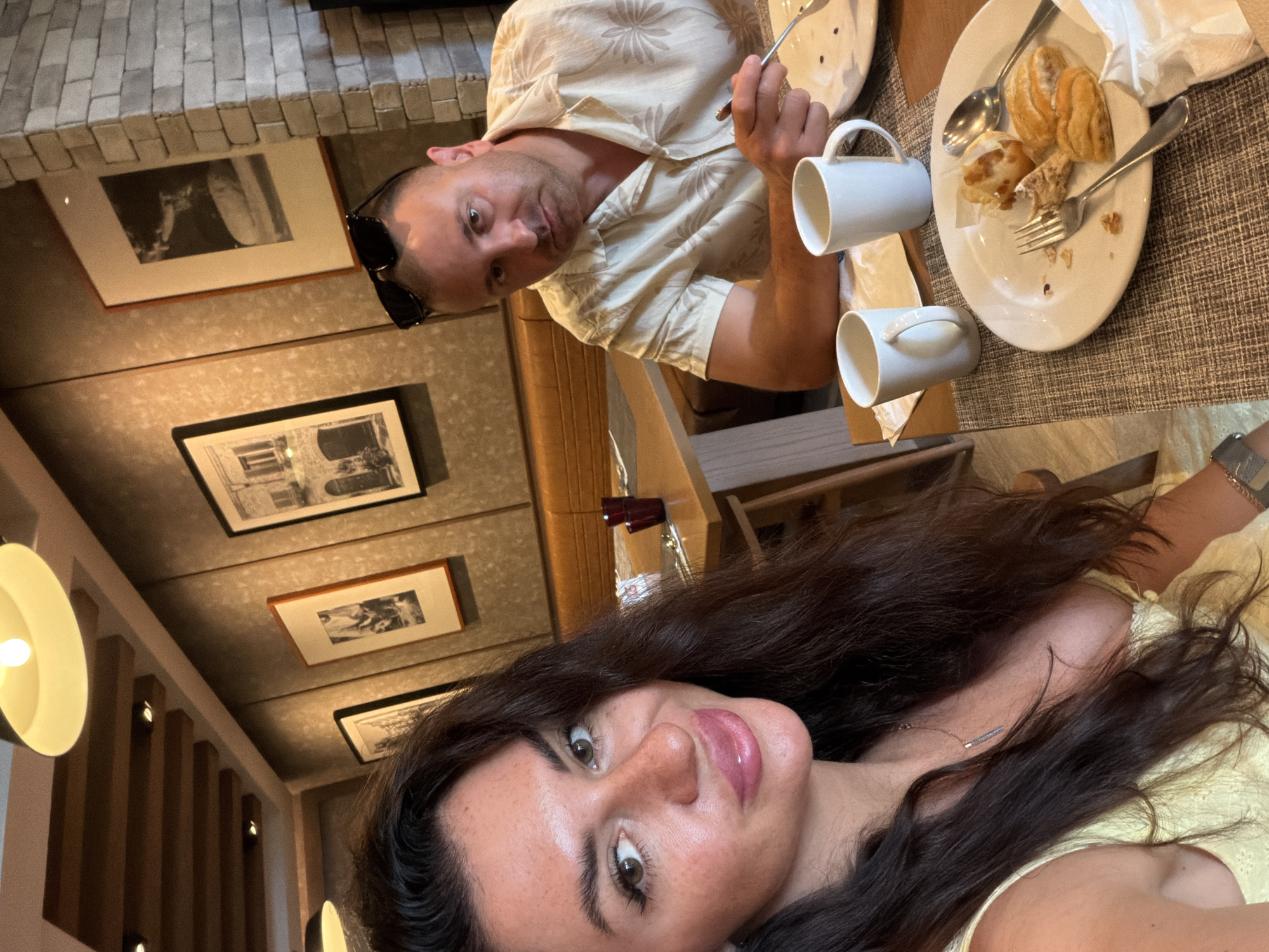 A man and a woman are seated at a wooden table in a restaurant. The man, wearing a patterned shirt and sunglasses perched on his head, is looking directly at the camera, holding a fork. In front of him are two white mugs and a plate with partially eaten pastries. The woman, with long dark hair and wearing a yellow dress, is also looking at the camera, with her face in the foreground. The background shows framed artwork on a textured wall, a brick column, and a leather banquette. The overall impression is a casual meal in an indoor dining setting. The location is stated to be Bang Muang, Thailand.