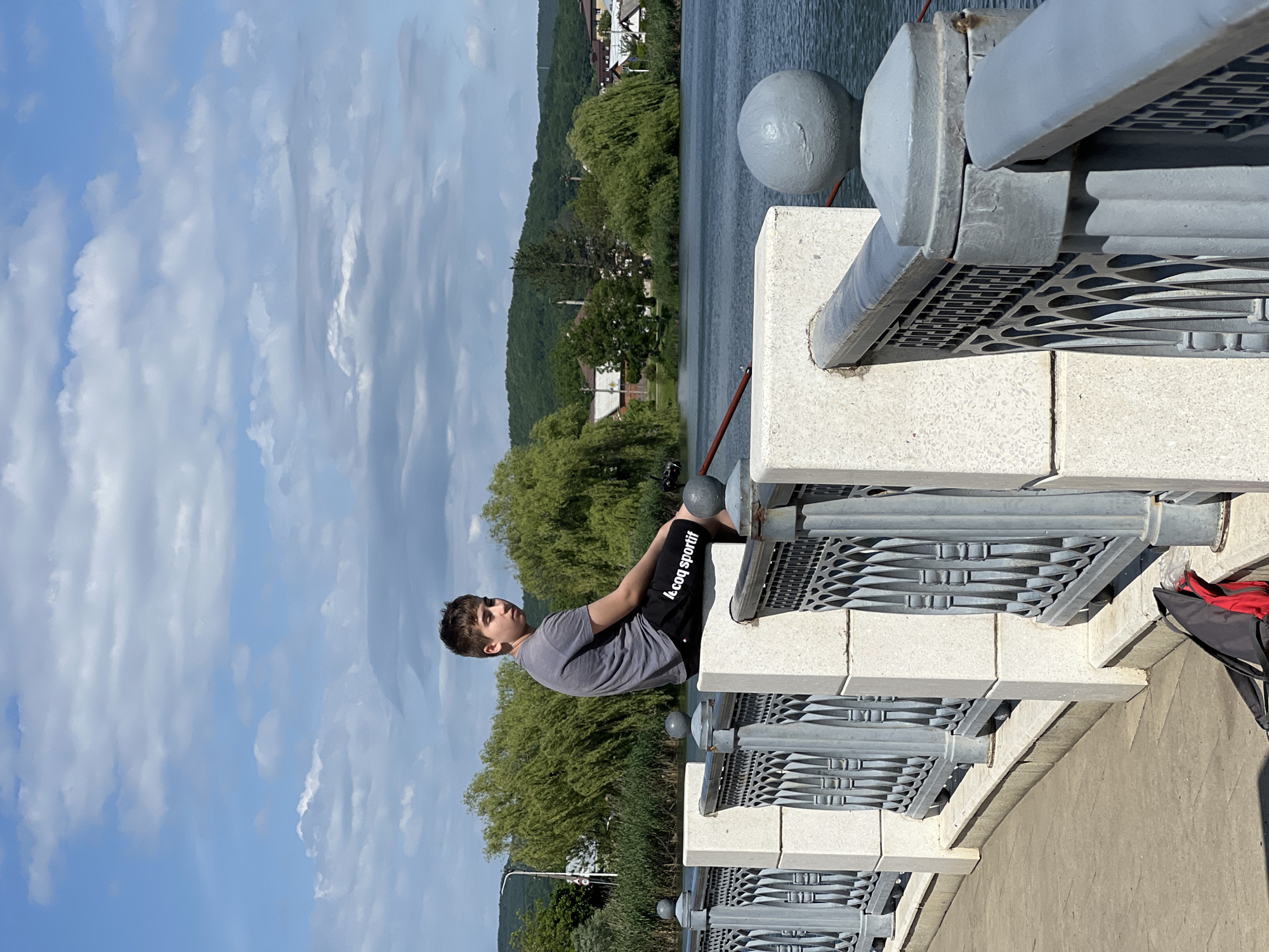 A young male sits on a gray bridge railing in Goryachiy Klyuch, Russia. He wears a gray t-shirt and black shorts with "Le coq sportif" printed in white. The bridge railing is composed of gray metal and white stone elements. Behind him is a view of green trees, hills, and a river or lake. The sky above is blue with scattered white clouds. Part of a red and gray bag is visible on the right side of the frame.