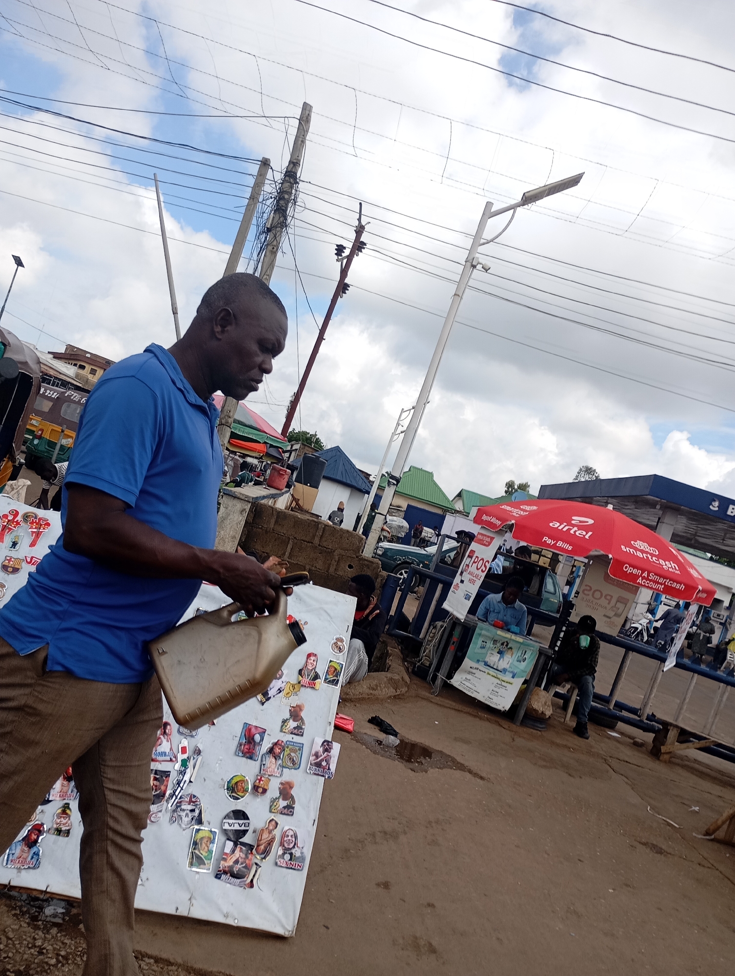 An outdoor scene in Gura Top, Nigeria, shows a Black man in a blue polo shirt and brown pants holding a tan plastic jerrycan with his right hand. He is looking down and to his right. In front of him is a white board covered in numerous colorful stickers, some depicting faces and logos. The background features a street with a blue barrier, a red umbrella with "Airtel" and "smartCash" logos, a POS machine, several people, and various buildings. Overhead, a complex network of power lines crisscrosses against a cloudy sky. The environment appears to be a bustling urban area with commercial activity.