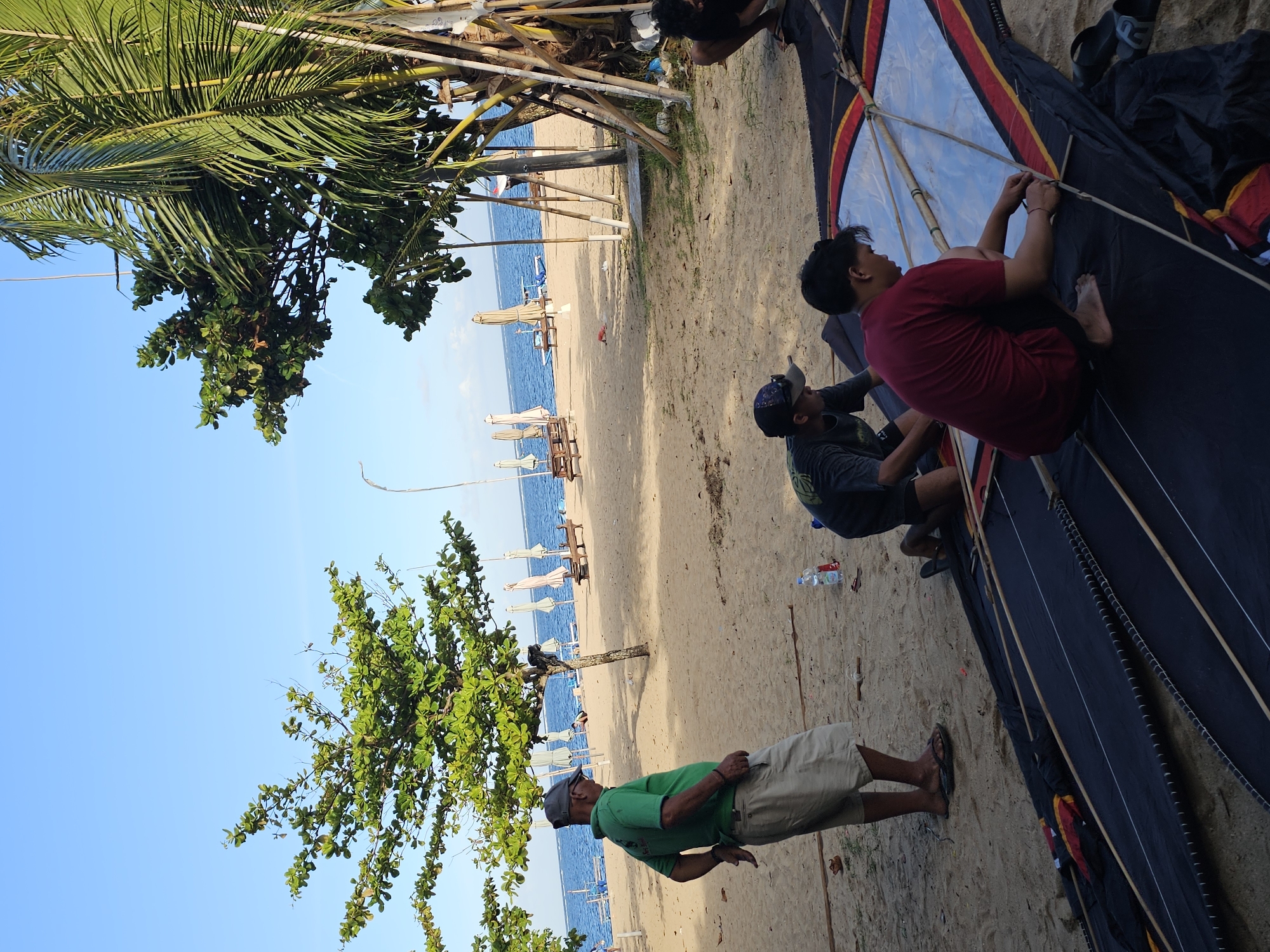 The image depicts a beach scene in Denpasar, Indonesia. In the foreground, two young men are working on a large kite with a black and white sail. One man is wearing a red shirt and the other is wearing a dark green shirt and a baseball cap. An older man wearing a green shirt, khaki shorts, a cap, and flip flops stands nearby, possibly observing. The beach is sandy, with trees providing shade. In the background, the ocean is visible, along with beach umbrellas and lounge chairs.