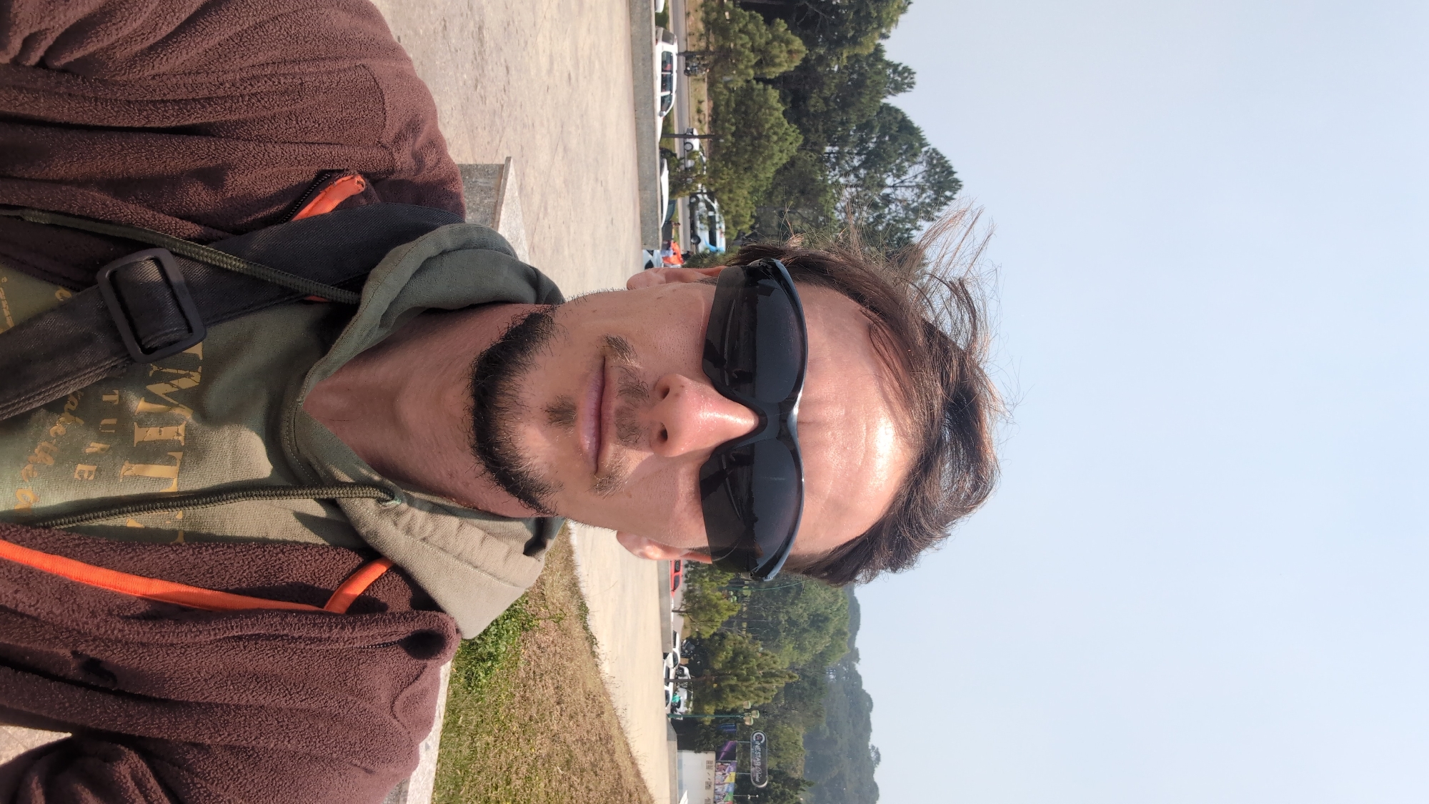 A man is facing the camera, with his head slightly tilted to the left. He is wearing sunglasses, a brown jacket, and a green shirt. He has a beard and mustache. The background is a paved area with trees and hills in the distance. There are cars and other vehicles parked along the paved area. The scene is a portion of the cityscape of Da Lat, Vietnam, where the man is presumably located.