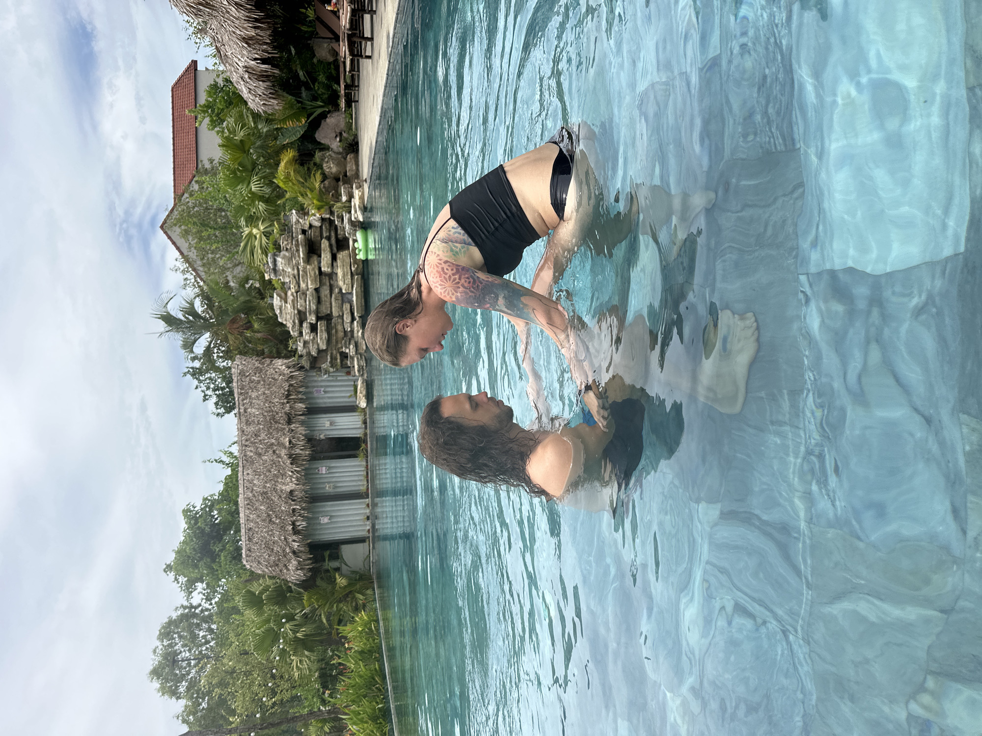 Two people are in a swimming pool in Nam Hoa Lư Ward, Vietnam. The person on the left has long, dark hair and is wearing blue swim trunks. They are facing the person on the right, who has blonde hair and is wearing a black bikini. The person on the right has tattoos on their arms and torso. They are reaching out to the person on the left. The water is clear and blue, with light reflecting off the surface. In the background, there is a building with a thatched roof, palm trees, and a rock formation. The sky is cloudy.