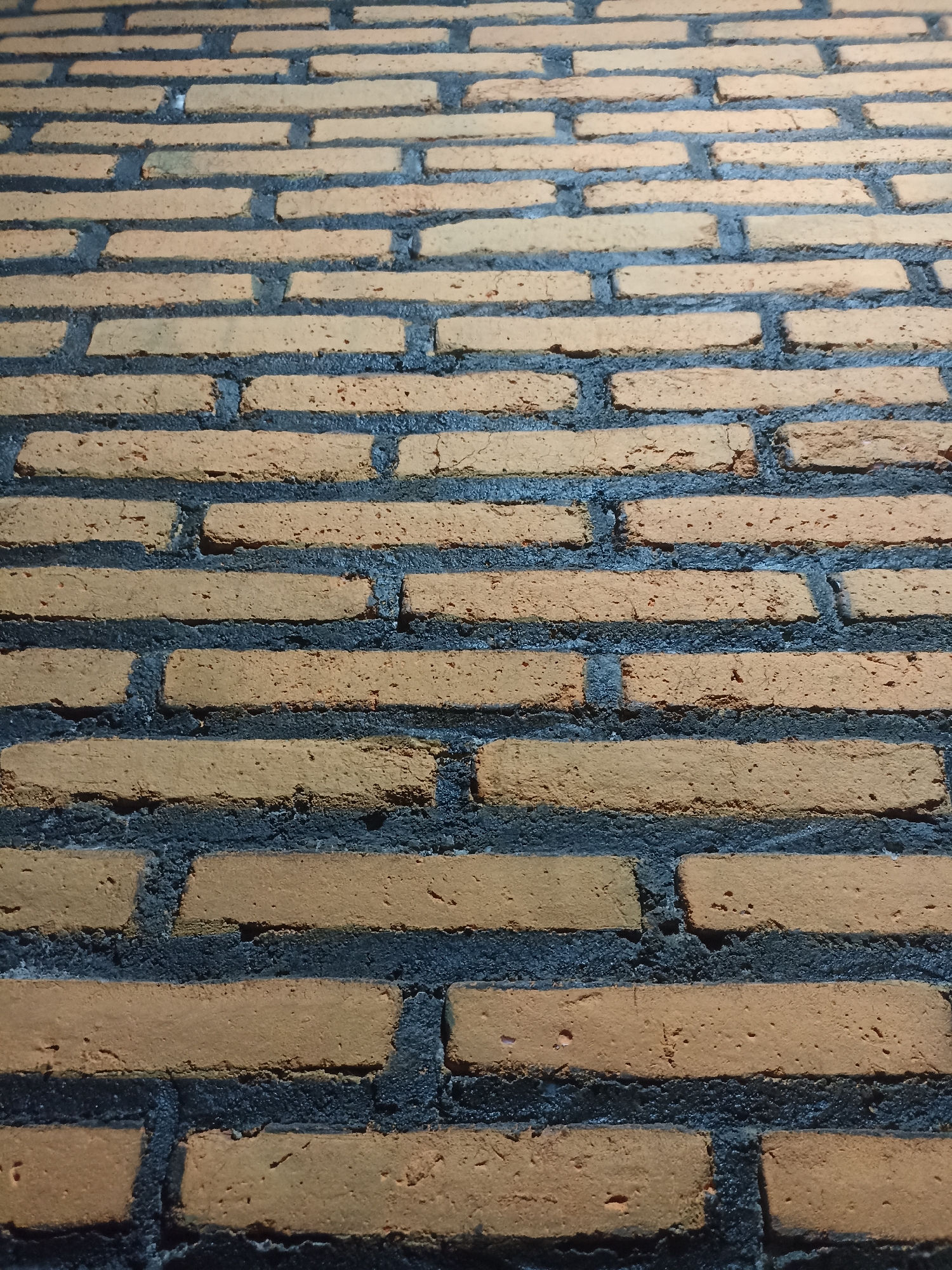 A brick wall with a consistent pattern of light brown bricks and dark gray mortar is visible. The bricks are laid in horizontal courses with staggered vertical joints. The perspective of the image suggests a slight upward angle, indicating the wall extends upwards and away from the viewer. The lighting appears to be artificial, casting shadows that emphasize the texture of the bricks and mortar. There are no people, animals, or specific objects present, and the scene is devoid of any discernible activity. The environment is solely the brick wall itself, with no surrounding context provided.