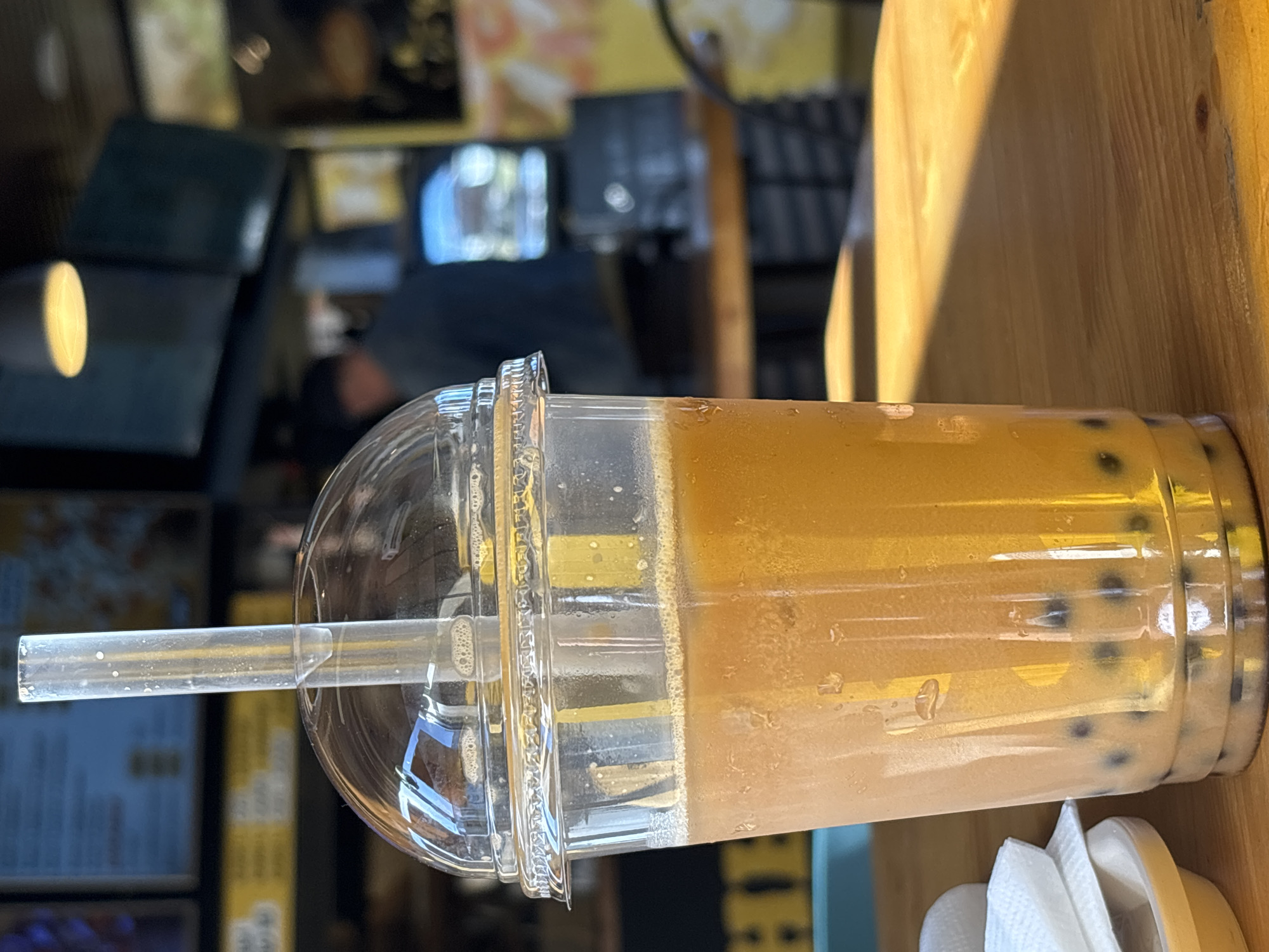 A plastic cup filled with a light brown and orange beverage, featuring tapioca pearls at the bottom, is positioned on a wooden table. A clear plastic straw extends from the domed lid of the cup. To the left of the cup, a small stack of white napkins is visible on a white saucer. The background is blurred, showing what appears to be an indoor setting with a counter, some televisions, and a person in the distance. The lighting suggests daylight, casting a warm glow on the table. The location is Unknown, Unknown.
