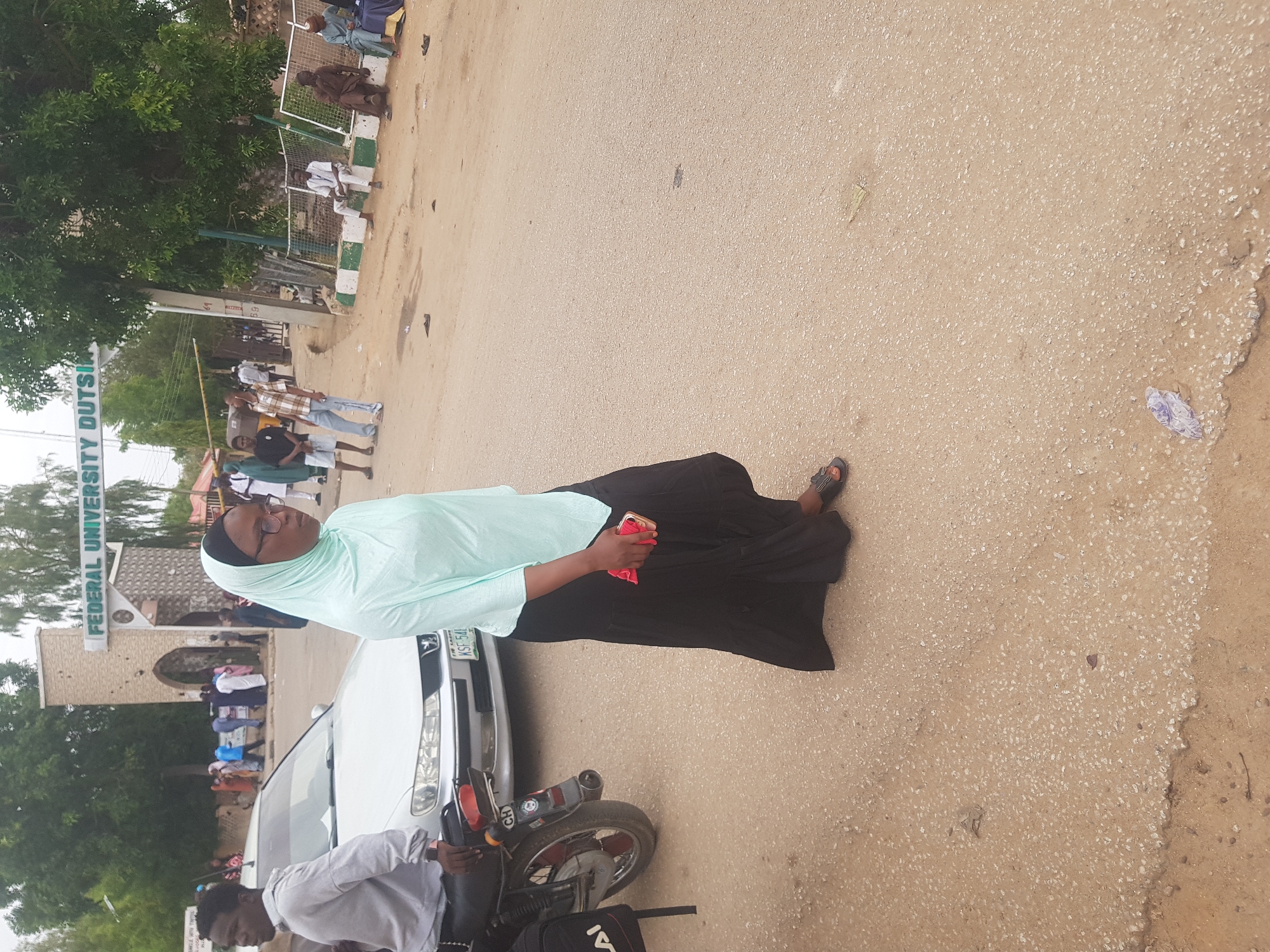 The image shows a woman in a light green hijab, light green shirt, and black skirt standing on a paved street in Dabawa, Nigeria, near the entrance to Federal University Dutse. She is holding a red object in her hand, possibly a phone or wallet. A white car is parked beside her, and a man is sitting on a motorcycle next to the car. In the background, other people can be seen walking near the university entrance. The weather appears to be overcast.