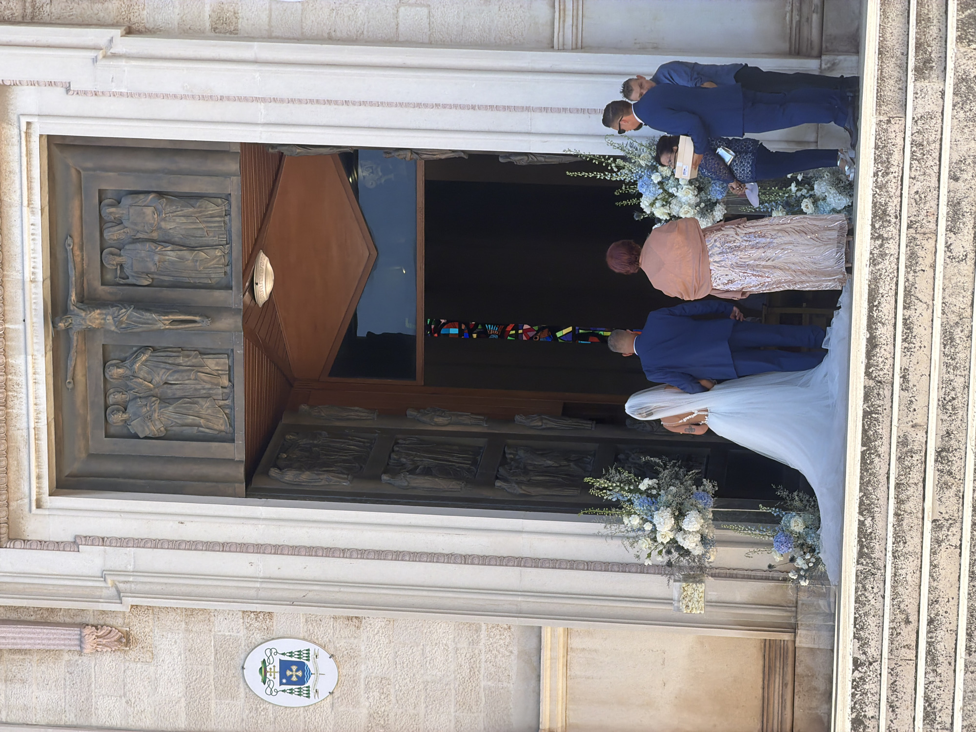 The image depicts a wedding ceremony at the Basilica of Saint Nicholas in Bari, Italy. The bride, wearing a white gown and veil, stands on the steps leading into the church. She is accompanied by the groom, dressed in a navy blue suit. Several other individuals are present, including a woman in a shimmering pink dress, a man in a navy suit holding a box, and another man in a blue shirt. Floral arrangements adorn the entrance. The basilica's facade, featuring stone carvings and a prominent coat of arms, is visible. The scene is set on a bright, sunny day.
