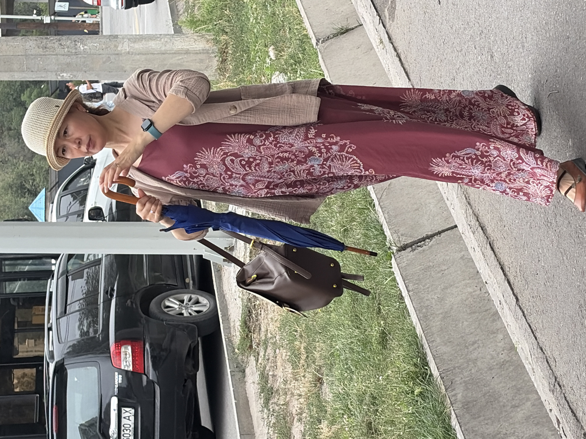 A woman with dark hair, wearing a beige straw hat, a beige cardigan, and a long maroon dress with a white paisley print, walks on a paved sidewalk next to a grassy area. She holds a closed blue umbrella in her right hand and a brown leather backpack hangs from her left shoulder. She is wearing brown flats with ankle straps. A black SUV is parked on the left side of the image. The scene appears to be in an urban environment.