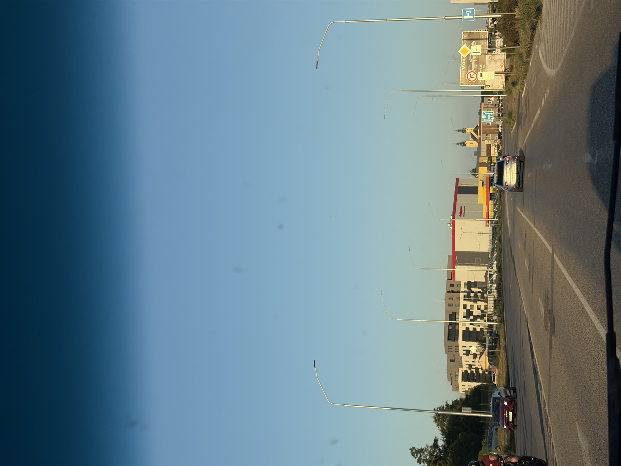 The image captures a view from a vehicle's dashboard, looking out onto a road in the city of Nitra, Slovakia. The sky is clear and blue, with the sun casting long shadows, indicating it is likely late afternoon or early evening.
In the foreground, the asphalt road is visible with lane markings. A silver car is seen driving away from the camera in the right lane. To the left, a red car is approaching the camera in the left lane. A portion of a motorcycle and its rider is visible on the far left edge of the frame.
The middle ground features a modern, multi-story building with a distinctive facade of white and dark grey geometric patterns. Adjacent to this building is a large commercial structure with a red roof, possibly a supermarket or retail store, indicated by signage that partially reads "SHOP". Further in the distance, a yellow church with two prominent spires stands out against the skyline.
The right side of the road is lined with tall streetlights and a billboard with various advertisements and signs, including a directional sign for "NITRA". There are also some smaller vehicles parked along the side of the road and grassy areas with bushes. The overall environment appears to be a suburban or commercial area of the city.