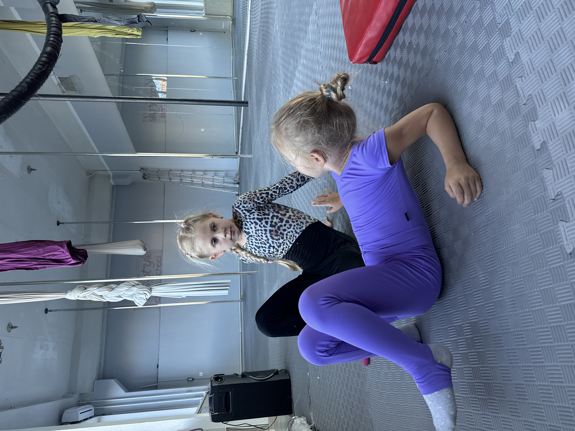 Two young girls are sitting on a grey, textured floor, possibly in a dance or gymnastics studio. The girl on the left is wearing a leopard-print top and black leggings, with her hair in a braid. She is looking towards the camera. The girl on the right is wearing a vibrant purple leotard and leggings, with her hair tied up in a bun. She is facing away from the camera and appears to be reaching out. In the background, there are aerial silks hanging from the ceiling, and a red object that looks like a mat or prop. There are also metal poles and what appear to be mirrors on the walls. A black box, possibly a speaker or equipment, is visible on the floor. The overall impression is a training or practice session. There is some faint text visible on the wall in the background, which appears to read "style school". The lighting suggests it is daytime, and the indoor setting indicates no specific weather conditions.