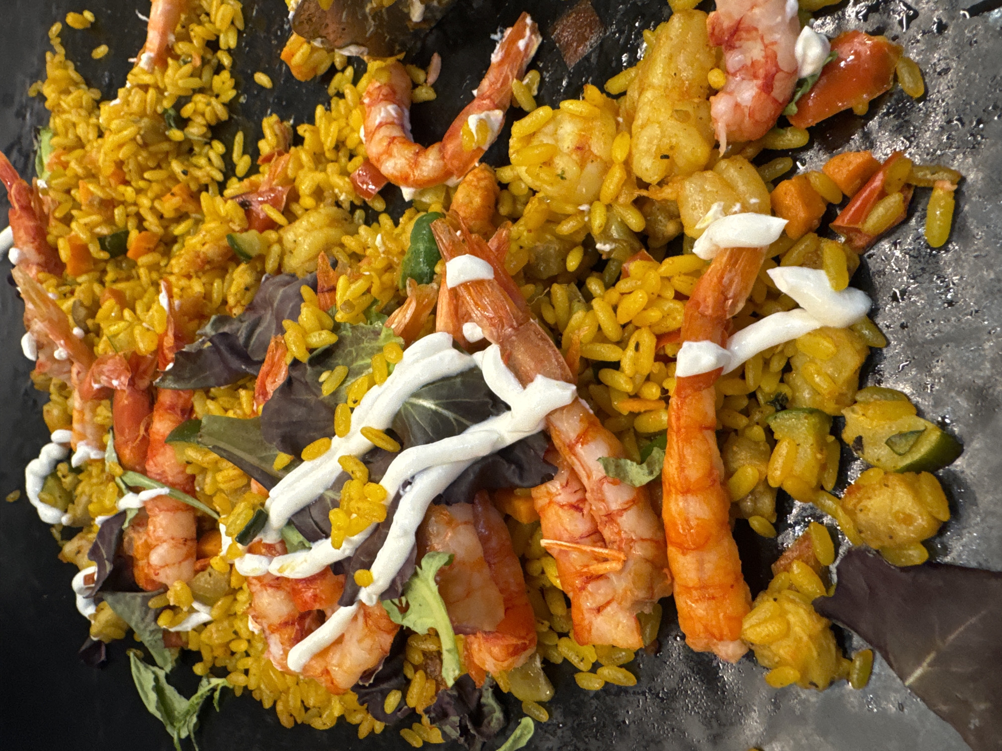 The image shows a close-up view of a dish of paella on a dark, flat surface, possibly a table. The paella consists of yellow rice, pieces of shrimp, and some green and red vegetables, as well as a drizzle of white sauce. There are also some dark leafy greens mixed in. The dish is artfully arranged, with the shrimp prominently displayed on top. The lighting is bright, casting some shadows on the food. The focus is on the food itself, with the background being dark and out of focus.