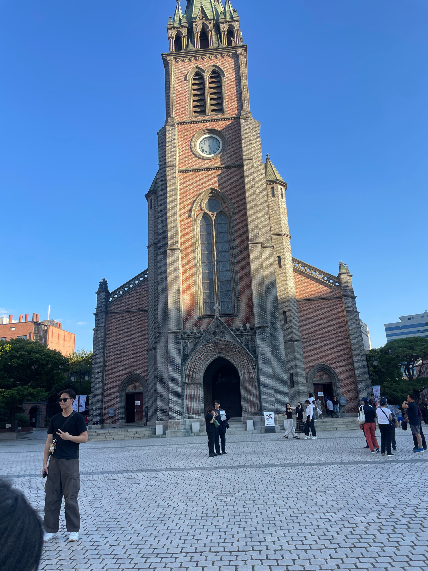 The image depicts Myeongdong Cathedral in Seoul, South Korea, on a clear, sunny day. The grand brick cathedral dominates the background with its tall bell tower, featuring arched windows and a clock face. The foreground is paved with cobblestones, and several individuals are visible.
In the left-center of the frame, a young man wearing sunglasses, a black t-shirt, and loose-fitting olive-colored pants stands facing left, holding a phone in his left hand and making a peace sign with his right hand. He is wearing white sneakers and has a small, light-colored bag hanging from his right shoulder.
To the right of the main entrance of the cathedral, a woman in black is holding a white sign. Further to the right, a group of people are gathered, some walking and others standing. One individual wears a white top and a blue baseball cap, while another is dressed in a dark top and lighter pants. A man in a black t-shirt and dark shorts is also present, as is a person wearing a dark top and red pants.
The environment appears to be a public square or plaza in front of the cathedral. Trees and other buildings are visible in the background. The overall impression is of a serene, daytime scene at a prominent landmark.
