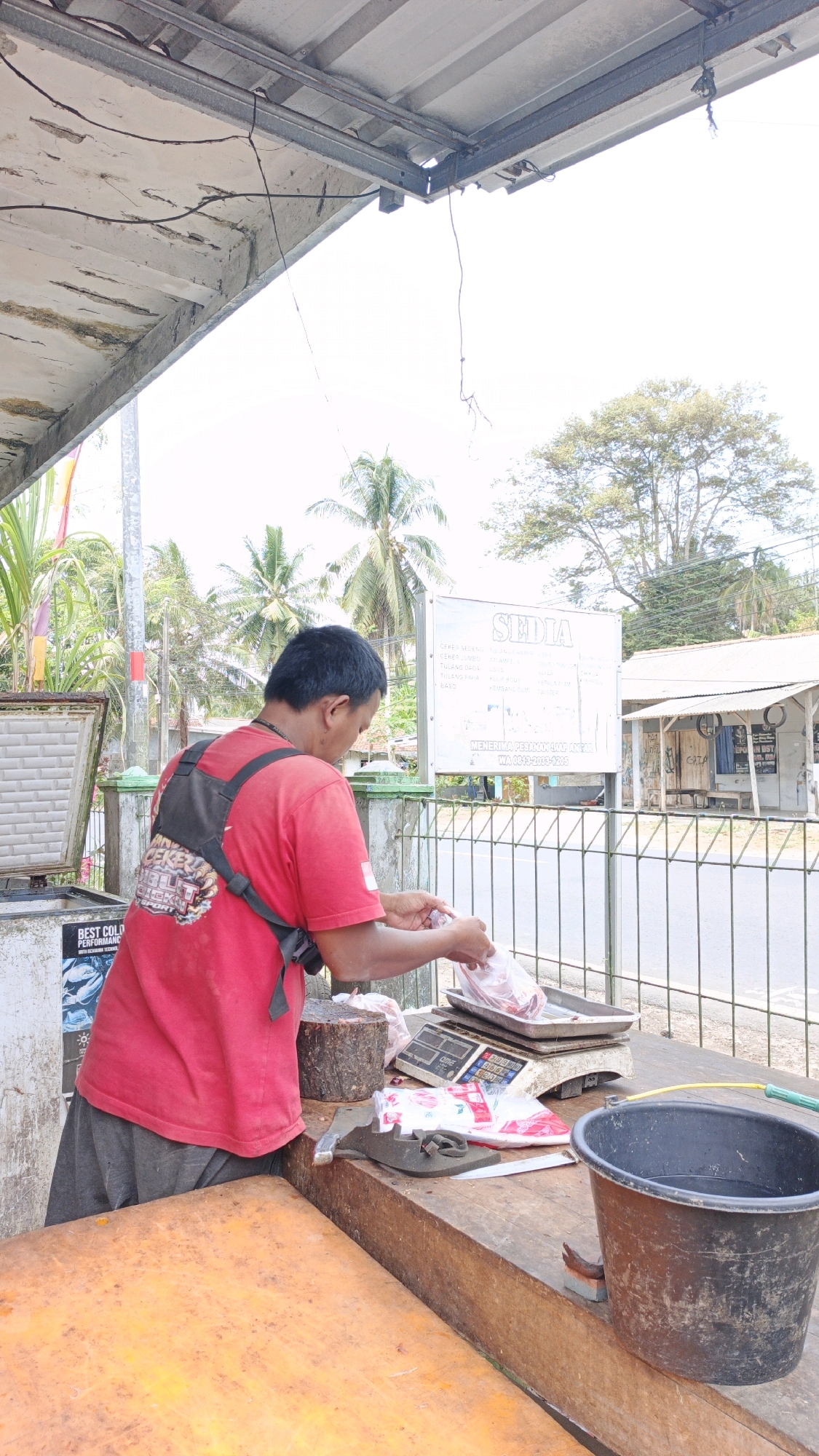 An outdoor butcher's stall is depicted in Cibenda, Indonesia, under a corrugated metal roof supported by a metal frame. A man wearing a red shirt and dark shorts stands behind a wooden counter. He is weighing meat in a plastic bag on a digital scale. To his left, a chest freezer is partially visible, with a label reading "BEST COLD PERFORMANCE." On the counter, a wooden chopping block, a knife, and a black bucket filled with water are present. In the background, a white sign with Indonesian text, palm trees, and a small building with a fence suggest a rural or semi-urban setting. The overall scene captures a moment of commerce in a typical Indonesian marketplace.