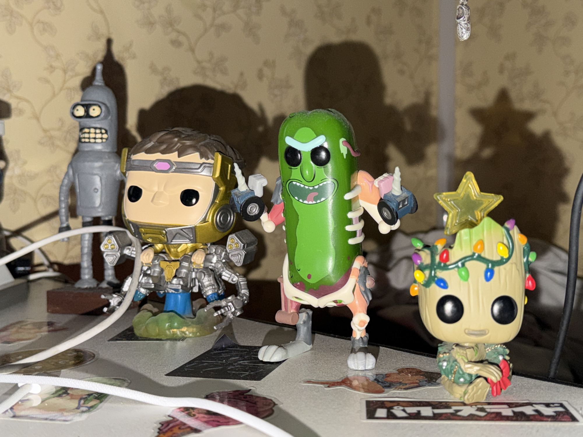 A collection of four Funko Pop figures are displayed on a surface. From left to right, the figures include Bender from Futurama, a golden and silver armored figure, a green pickle figure with a maniacal grin, and a Groot figure adorned with Christmas lights and a star. The background features a textured, patterned wallpaper and shadows cast by the figures. White cords are visible in the foreground. The location is described as Unknown, Unknown.