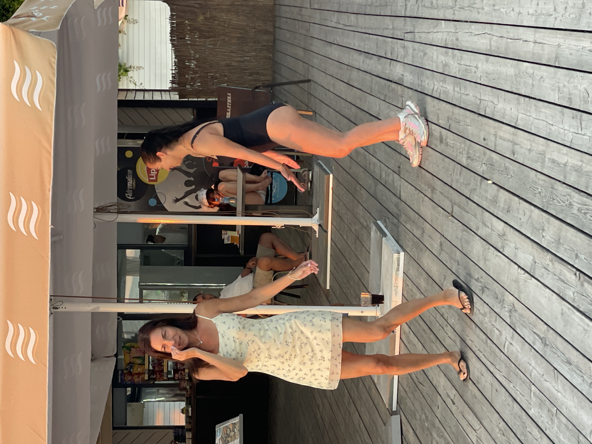 A full shot captures two young adult women dancing on a wooden boardwalk in Sochi, Russia. The woman on the left, wearing a light-colored, floral-print dress and black flip-flops, has her left hand to her face. The woman on the right, dressed in a black one-piece swimsuit and pink and white sneakers, is in mid-dance pose with her arms extended. Behind them, a thatched barrier and a building with a sign featuring a silhouette of people dancing and "Adrenaline Rush" visible. The boardwalk is laid with weathered gray wooden planks, and a large, tan umbrella with wavy white patterns is overhead. In the background, other people are seated at tables under a canopy, and a man in a white shirt and shorts is visible. The lighting suggests it is late afternoon or early evening.