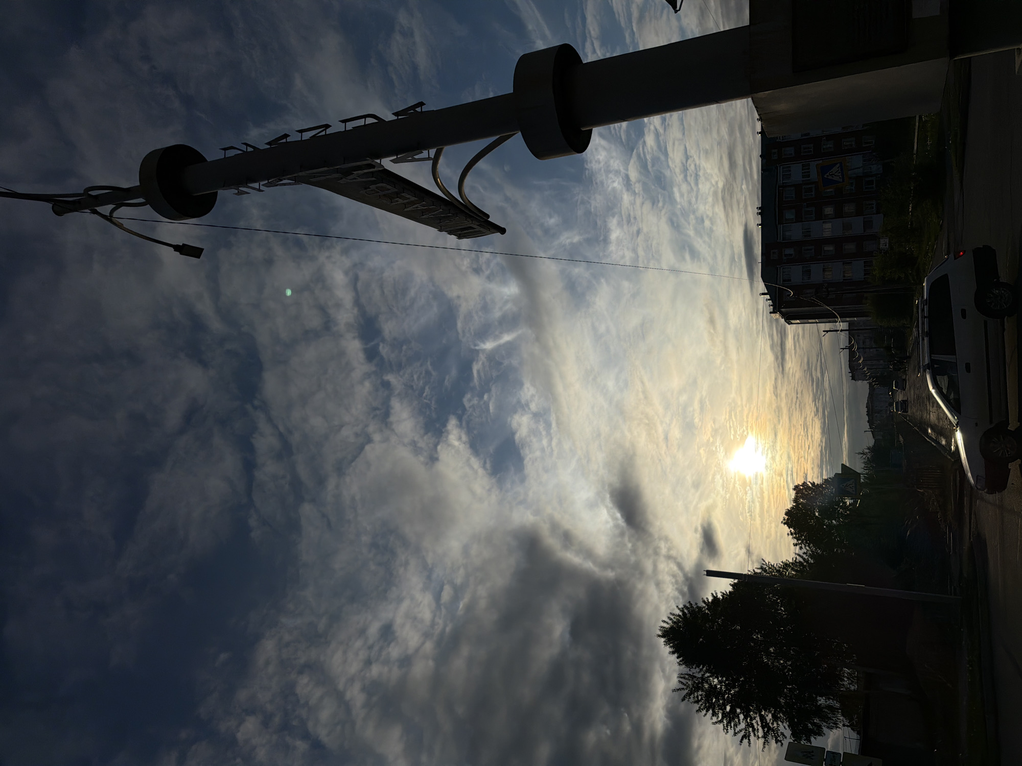 The image depicts an urban street scene at either sunrise or sunset, characterized by strong backlighting from a low sun. The location is identified as Shpaleva Street (Улица Шпалева) in Birobidzhan, Russia, indicated by Cyrillic text on a street sign.
In the foreground, on the right, a large, dark-silhouetted structural pole or monument stands vertically. A horizontal sign, also silhouetted, extends from this pole to the left, displaying partial Cyrillic text, reading "ШПАЛЕВА" (Shpaleva). A bright green lens flare is visible in the upper mid-left quadrant of the sky. On the left foreground, several trees are silhouetted, appearing dark against the brighter sky, with a dark utility pole partially visible behind them.
A paved road extends from the lower center towards the background. A light-colored (likely silver or white) station wagon or sedan is positioned in the lower mid-ground, driving from left to right, with its front end illuminated by the sun's glare. Multiple other vehicles are visible further down the road, receding into the distance. Two blue and white pedestrian crossing signs are discernible: one on the left side of the road near the silhouetted trees and another further down near the buildings on the right.
In the mid-background and background, multi-story residential buildings are visible on both sides of the street. The most prominent building, located on the right side of the road, is a large, dark brick or panel structure with numerous windows, consistent with Soviet-era architecture. The sky above is partially cloudy, with darker, denser clouds near the top left and thinner, more diffuse clouds allowing the sun's rays to penetrate and illuminate the horizon line. No individuals are clearly discernible.