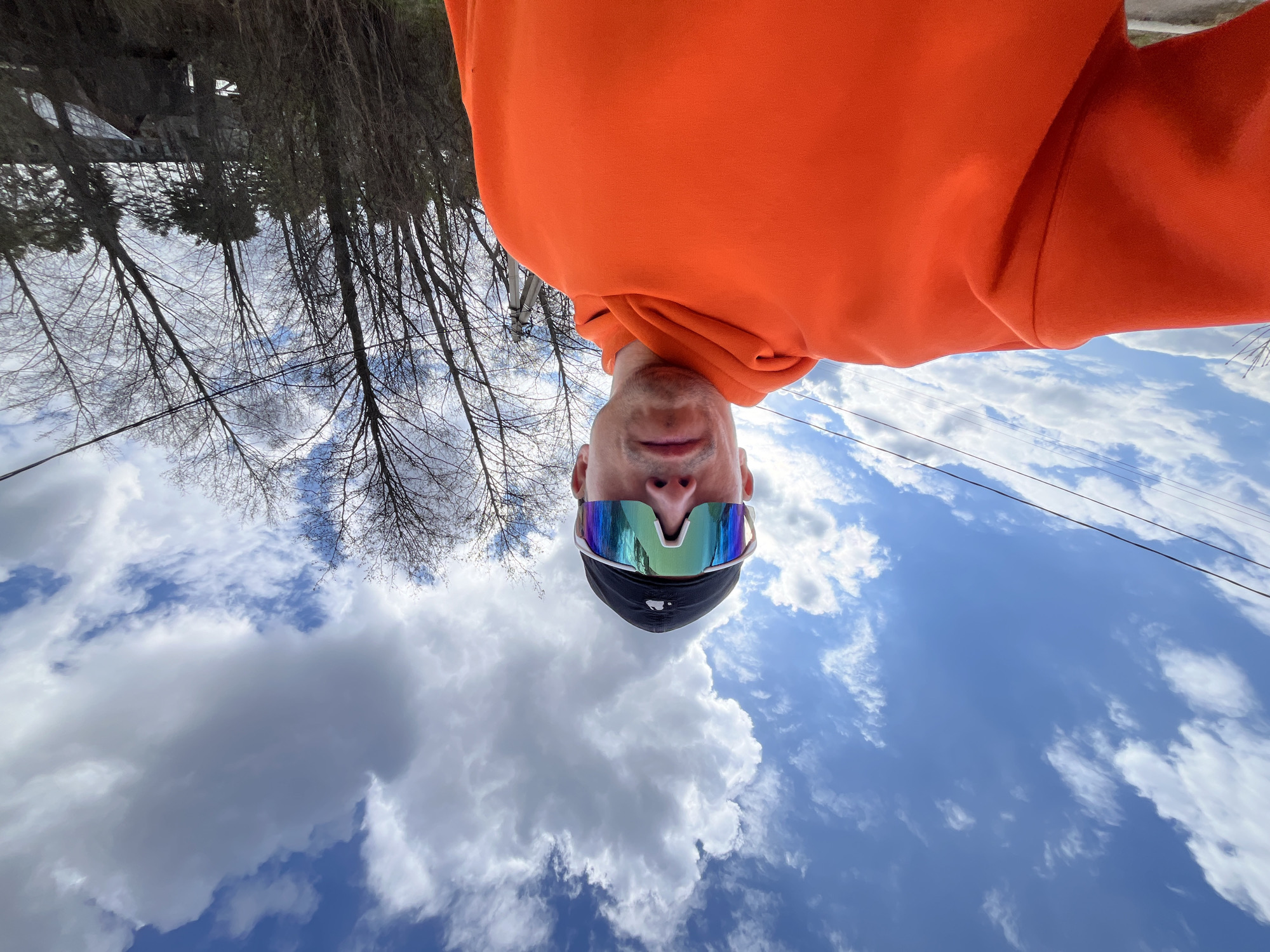 This image is an outdoor portrait, likely a selfie, taken from a low angle looking upwards, resulting in the subject appearing inverted. A person, whose lower face (chin, mouth, nose) is visible, is the central subject. They are wearing a vibrant orange hooded sweatshirt and large, modern sports sunglasses with highly reflective, iridescent lenses that show hues of blue, green, and purple. A dark-colored cap or beanie is also visible beneath the sunglasses.
The background consists of a bright blue sky with numerous white, fluffy cumulus clouds, indicating clear and sunny daytime weather. On the left side and top of the frame, there are many bare trees with intricate branch patterns, suggesting late winter or early spring. Some faint, thin power lines or utility wires crisscross the sky in the upper right quadrant.
The image captures a moment in what appears to be an outdoor setting in Chaikovsky, Russia, on a bright day. The person's expression is neutral, partially obscured by the sunglasses and the inverted perspective. There is no visible text in the image.