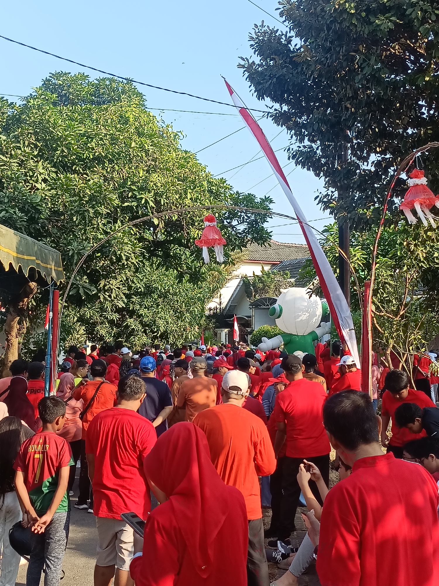 This image depicts an outdoor gathering of people, likely in a residential area of Kediri City, Indonesia, given the context provided. A large crowd, predominantly wearing red shirts, is visible, suggesting a communal event or celebration. The atmosphere appears festive, with decorative elements like red and white flags and hanging red paper ornaments adding to the ambiance. Trees and houses form the backdrop, indicating a suburban or semi-urban setting. Some individuals are engaged in activities such as taking photographs with their mobile phones. The presence of a large inflatable green and white character further hints at a celebratory occasion, possibly a festival or local fair. The scene is captured under a clear blue sky, suggesting it is daytime.