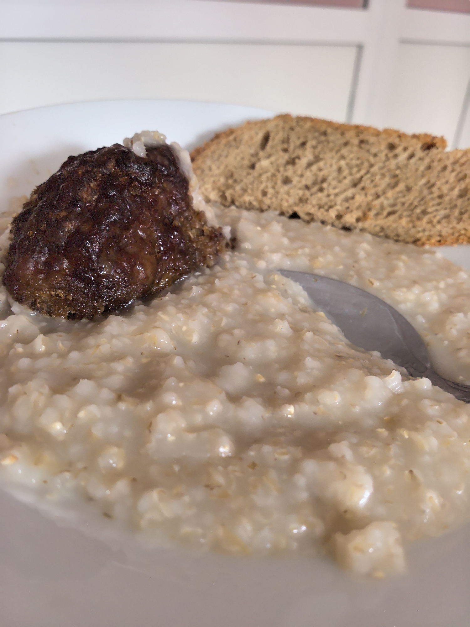 A plate of food is shown. The food consists of oatmeal, a meat patty, and a slice of brown bread. A spoon rests in the oatmeal. The location appears to be indoors, possibly a kitchen or dining area, given the white background with window frames. The city and country are unknown.