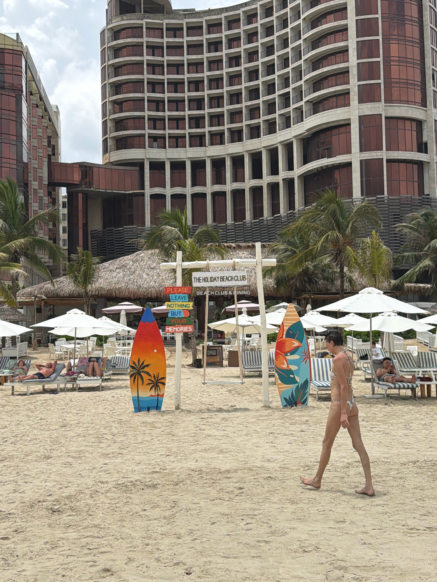 A wide shot of a beach scene in Da Nang, Vietnam, features a large, modern hotel complex in the background with tan and reddish-brown facades and numerous windows. In the foreground, a sandy beach is dotted with lounge chairs and umbrellas. Several people are visible, including a woman in a bikini walking across the sand towards the right side of the frame. A surfboard with a sunset and palm tree design stands vertically on the left side of the image. A directional sign with humorous beach-related phrases like "Please Leave Nothing But Your Memories" is also present. Palm trees and thatched-roof structures suggest a tropical resort environment. The sky is overcast.