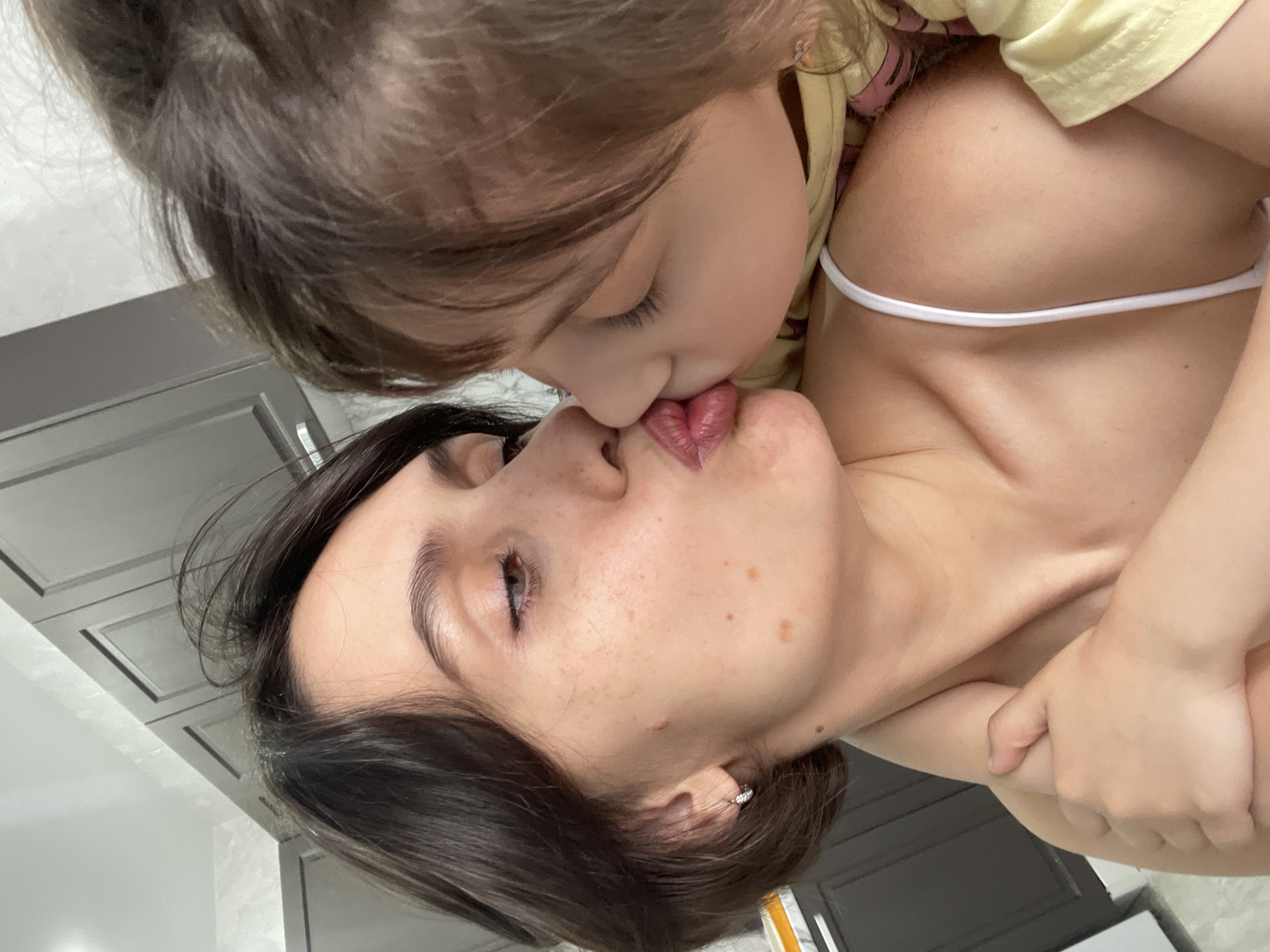 A mother and child are kissing in an indoor setting. The woman has fair skin, short dark hair, freckles, and is wearing a white spaghetti strap top. The child, with fair skin and brown hair, is wearing a yellow shirt. They are in front of a wall or cabinet with a gray tile pattern. The image does not provide sufficient information to determine if the location is in Izhevsk, Russia.