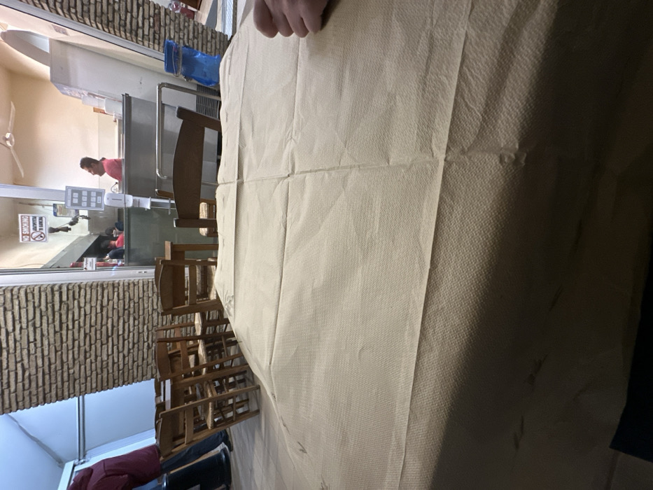 This vertical indoor photograph, taken in Nea Erithrea, Greece, depicts a scene within what appears to be a restaurant or cafe. The foreground is dominated by a light beige or off-white paper tablecloth covering a table, exhibiting visible folds and a subtle, ridged texture. A person's arm and hand are visible in the upper right corner, resting on this tablecloth.
In the mid-ground, a stack of several wooden chairs is neatly arranged, hinting that the establishment might be closed, preparing for service, or undergoing cleaning. To the left, a textured wall, possibly made of brick or stone, adds architectural detail to the space.
The background features a service area behind a clear glass partition. A person wearing a pink or red shirt is visible leaning over a stainless steel counter, engaged in an activity that suggests work, possibly food preparation or service. A blue container or bag and some white dispensers are also present on the counter. Through the glass, in the deeper background, other individuals are partially visible, seated in what appears to be another section of the eatery. A white ceiling fan hangs in the upper left background.
A distinct "NO SMOKING" sign, written in Greek ("ΑΠΑΓΟΡΕΥΕΤΑΙ ΤΟ ΚΑΠΝΙΣΜΑ"), is affixed to the glass partition. The lighting is ambient, suggesting a regular time of day without strong, direct sunlight. The overall atmosphere conveyed is one of a quiet, functional space.