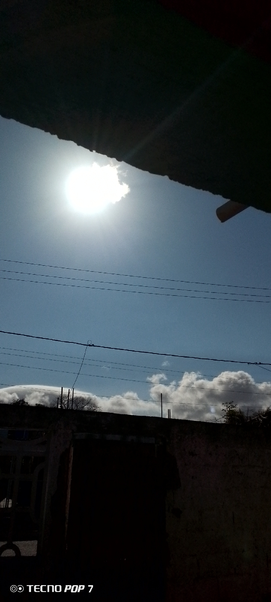 The image displays an outdoor scene dominated by a bright, sunny sky. A brilliant, almost-white sun is prominently featured in the upper left section, partially veiled by a white cloud, casting intense light across the frame.
Several dark utility or power lines traverse horizontally through the middle of the sky. Below these lines, the silhouettes of what appear to be rooftops or the upper parts of buildings are visible, with scattered white and grey clouds forming a lower layer on the horizon. Some dark, bare tree branches or structures can be discerned against the clouds in the middle ground.
The top and bottom sections of the image are obscured by large, dark, silhouetted structures, likely an overhang or roof at the top, and a wall or building in the foreground at the bottom. These elements are heavily underexposed, rendering specific details indistinguishable, though subtle textures of what might be concrete or brick can be seen in the bottom right crop. A faint orange pipe-like object is visible under the top overhang on the right side.
The time of day is clearly daytime, characterized by bright, direct sunlight. The weather is sunny with a clear blue sky, broken by a few clouds. The utility lines suggest an urban or suburban setting in Altagracia, Venezuela. No people are visible, and no specific activity is taking place.
In the bottom left corner, the text "TECNO POP 7" is visible, indicating the device used to capture the photograph.