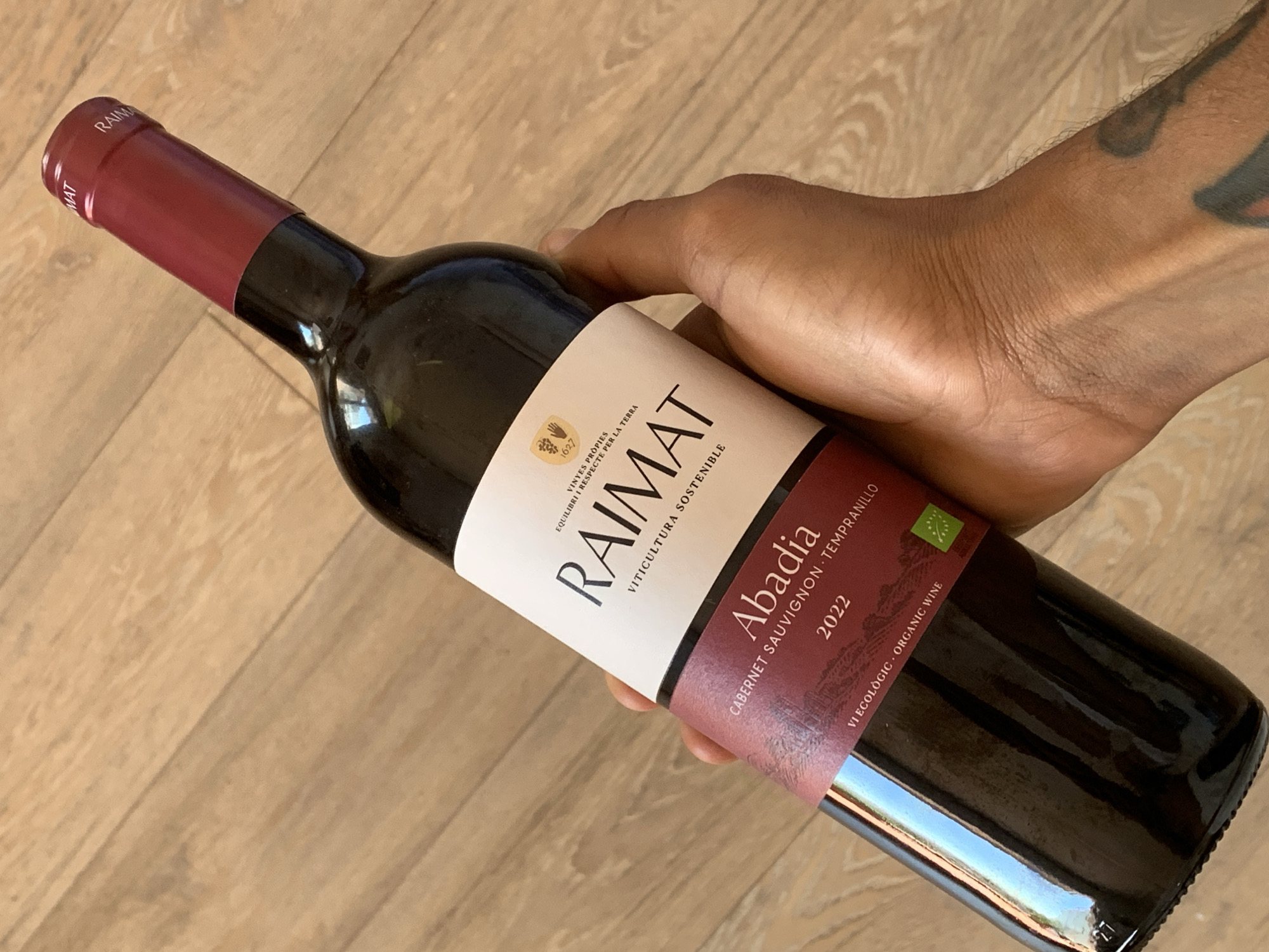 A hand holds a bottle of Raimat Abadia wine. The bottle is dark glass and has a dark red capsule on the neck. The label is white with black text and a dark red band at the bottom. The text on the label reads "RAIMAT VITICULTURA SOSTENIBLE", "Abadia", "CABERNET SAUVIGNON - TEMPRANILLO", and "2022 VI ECOLÒGIC ORGANIC WINE". The hand is holding the bottle at an angle against a light brown wooden floor. The image appears to have been taken indoors, possibly in a residential setting in Barcelona, Spain. The background is out of focus, showing only the wooden floor.