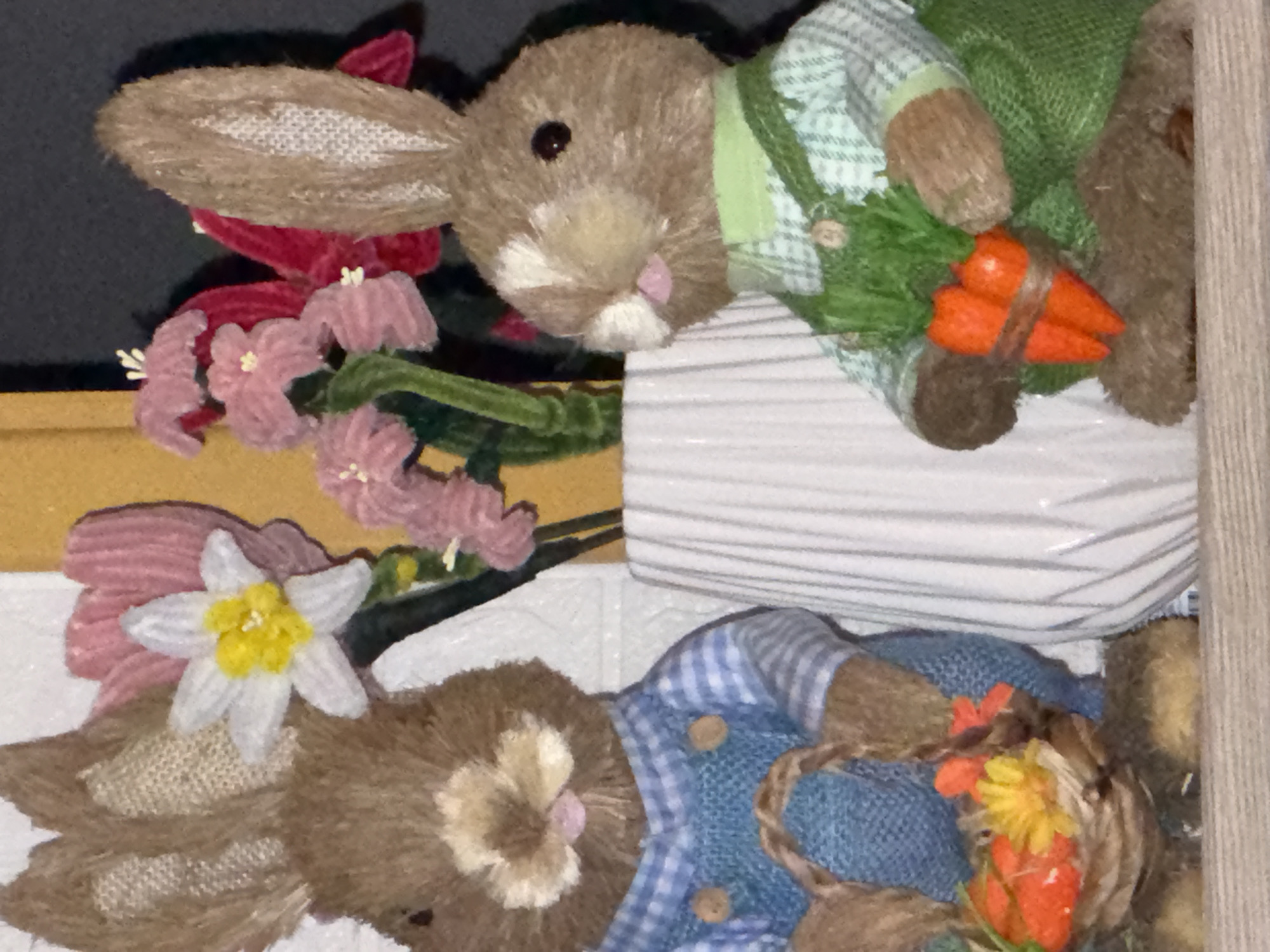 The image shows two stuffed toy rabbits. The top rabbit is lying on a white, striped surface, wearing a green and white checkered shirt, green overalls, and holding a bundle of carrots. The bottom rabbit is wearing a blue and white checkered shirt with blue overalls, and it is holding a basket of orange and yellow flowers. There are also artificial flowers, including pink blossoms and a white flower with a yellow center, around the rabbits. The background includes white and beige textured surfaces. The scene appears to be indoors, perhaps a display or a shelf. This image doesn't show any details that would indicate its specific location in Plovdiv, Bulgaria or any other place.