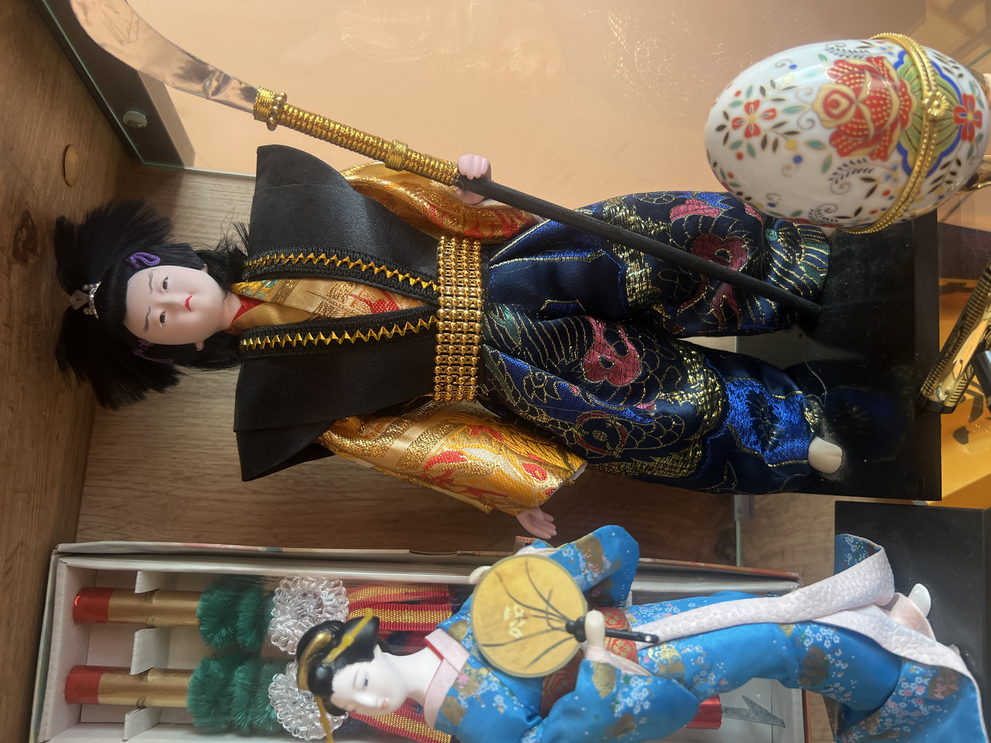 The image depicts two Japanese dolls and a decorative egg within a display case. The upper doll is dressed in a black, gold, and blue patterned robe and holds a glaive. The lower doll is dressed in a blue kimono and holds a fan. The decorative egg is white with floral patterns and gold detailing. The dolls appear to be in a private setting. There is no indication of Herakovo, Bulgaria.