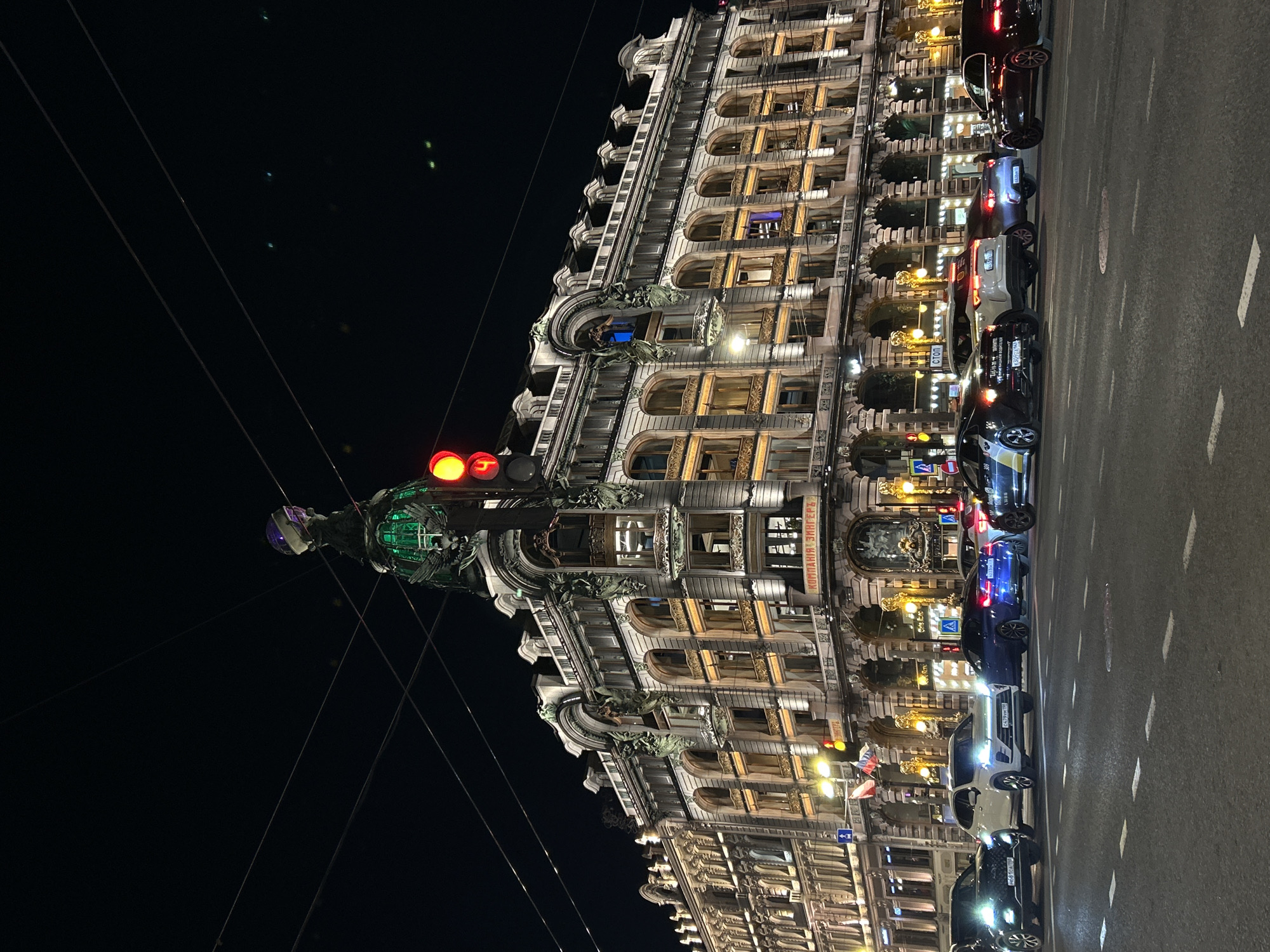 This media file captures a nighttime street scene in Saint Petersburg, Russia. A grand, ornate, multi-story building, likely historic, dominates the left side of the frame, its detailed facade beautifully illuminated. The architecture features numerous arched windows, balconies, intricate carvings, and pilasters, suggesting an Art Nouveau or similar elaborate style. Many of the windows show interior lights, and some feature small sculptures or decorative elements.
In the foreground, slightly to the left of center, stands a tall pole holding a traffic light displaying red signals. Below the traffic light is an elaborate, green-lit ornate structure, possibly a historic street lamp or a small monument, topped with a distinct purple-hued element. Several thin overhead wires stretch across the dark, clear night sky from this pole.
To the right of the building is a paved street with white lane markings. Several cars are parked along the curb, their headlights and taillights visible, adding to the nocturnal illumination. Two specific Russian license plates are partially visible on parked cars: "Р 629 МЕ 178" on a white SUV and "H 061 HA 178" on a black SUV. A blue street sign with a white arrow pointing right (indicating a one-way street or turn) is also visible near the parked vehicles.
Visible text on the building, specifically on a sign above the lower windows, reads "КОМПАНИЯ ЗИНГЕРЪ" (KOMPANIYA ZINGER'), which translates to "Singer Company," a key indicator of the building being the iconic Singer House (Dom Knigi). Another sign, "СТОП" (STOP), is also seen on the building facade. The overall scene is calm and static, with no people visible, emphasizing the illuminated architecture and the quiet night atmosphere of the city.