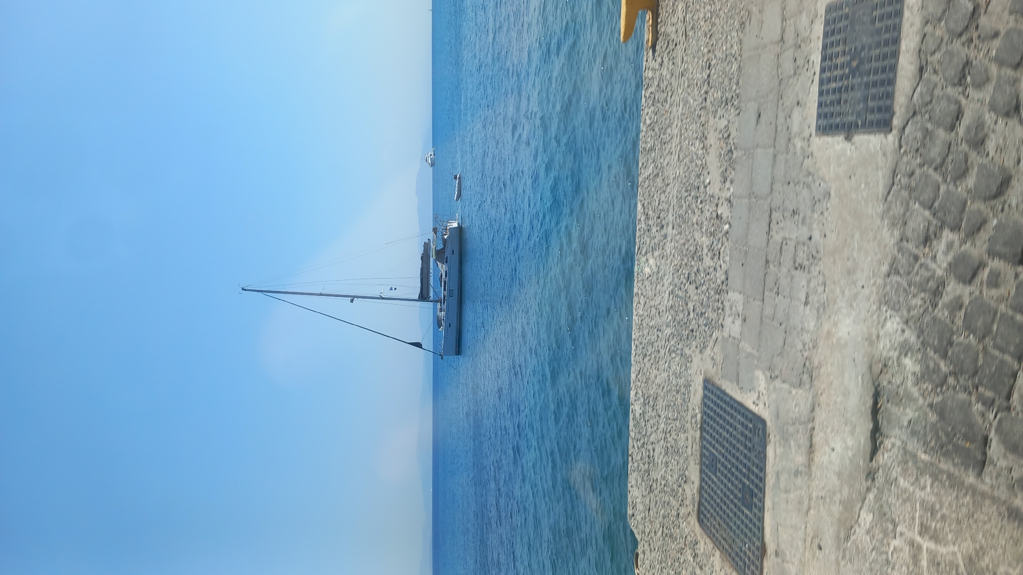A sailboat is moored in calm blue water near a paved waterfront in the city of Kos, Greece. The sailboat has a white hull and a dark sail. A small dinghy is tied to the stern of the sailboat. In the background, hazy mountains are visible on the horizon. The waterfront is made of grey cobblestones and concrete slabs, and features metal grates. A yellow cleat is visible on the right side of the frame. The sky is clear and blue.