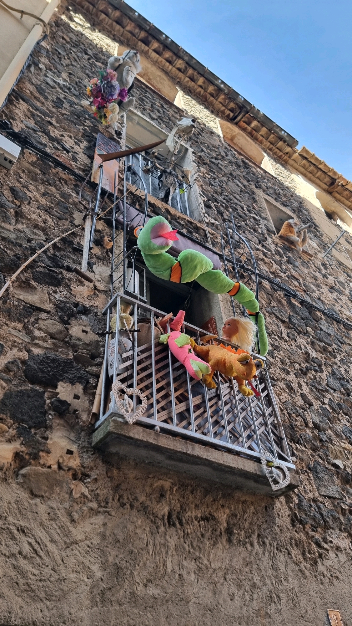 This is a low-angle, medium shot of stuffed animals placed on a balcony and exterior wall of a stone building in Castellfollit De La Roca, Spain. The building's facade is constructed of rough, dark stones, with some lighter-colored stucco visible. A narrow balcony, made of metal bars and a concrete ledge, juts out from the second story.
On the balcony, a large, green, segmented stuffed snake with an orange tongue is draped over the railing, extending upwards and to the right. Below it, a pink stuffed creature with a yellow tail and orange legs, possibly a frog or dragon, lies on its side. Next to it is an orange stuffed creature that resembles a dragon, also with a red tongue. A blonde doll head with blue eyes is also visible on the balcony. Decorative heart-shaped ornaments are attached to the balcony railing.
Above the balcony, another stuffed animal, resembling a grey, fluffy creature holding a bouquet of colorful artificial flowers, is affixed to the wall. Further up the wall, a white stuffed animal resembling a horse or skeleton is visible. The sky above is clear and blue. The overall scene appears to be an outdoor decoration.