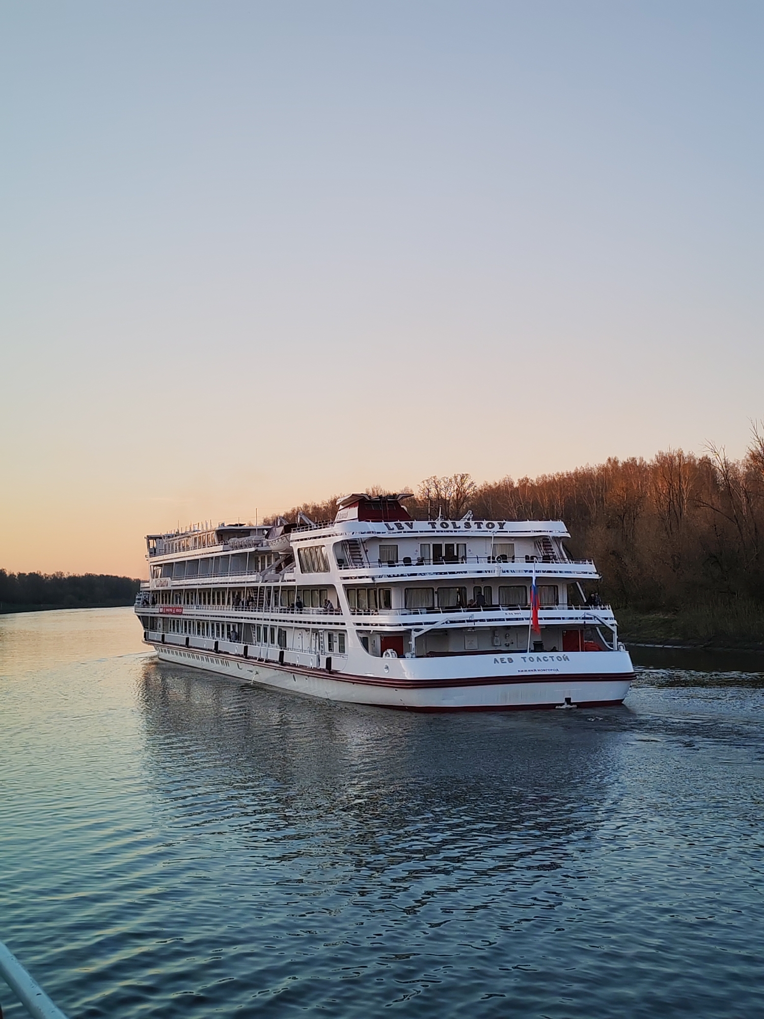 A four-deck river cruise ship, the "LEV TOLSTOY," with the words "ЛЕВ ТОЛСТОЙ Нижний Новгород" on its stern, sails on a river at dusk. The ship is white with a red stripe along its base and a red roof. The Russian flag flies at the rear. Trees line the river bank on the right. The sky is a pale mix of blue and orange from the setting sun. The location is likely near Yakhroma, Russia, given the ship's origin.