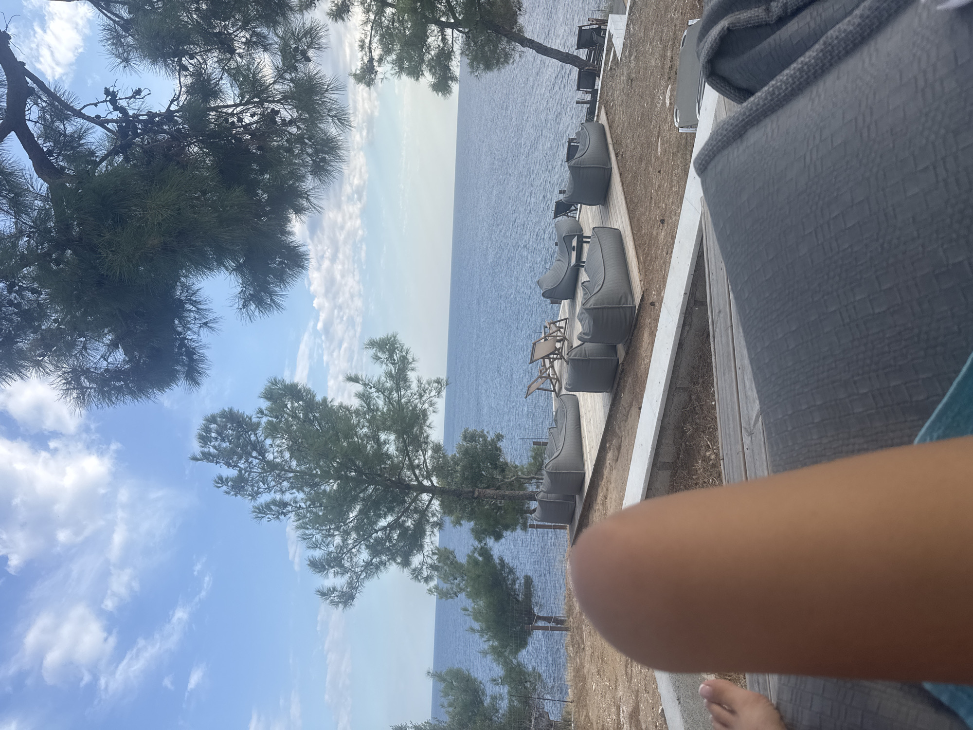 The image captures a serene outdoor setting with a view of the Aegean Sea, likely at a beach club or resort in Alyki, Greece. In the foreground, a person's leg and foot are visible, suggesting they are lounging or relaxing on a dark gray upholstered seating area. Beyond this, a wooden deck features several plush, gray beanbag-style chairs and low tables, creating a comfortable seating arrangement. A few other chairs are visible on the deck as well.
The middle ground showcases a shoreline with scattered trees, their green foliage contrasting with the sandy ground. A wire fence runs along the edge of the property. The sea itself is a deep blue, stretching to the horizon under a sky filled with scattered white clouds and patches of blue. Pine trees, characteristic of the Mediterranean landscape, frame the top of the image, their branches casting shadows and adding a natural element to the scene. The overall impression is one of tranquility and relaxation, with the natural beauty of the Greek coast as a backdrop.