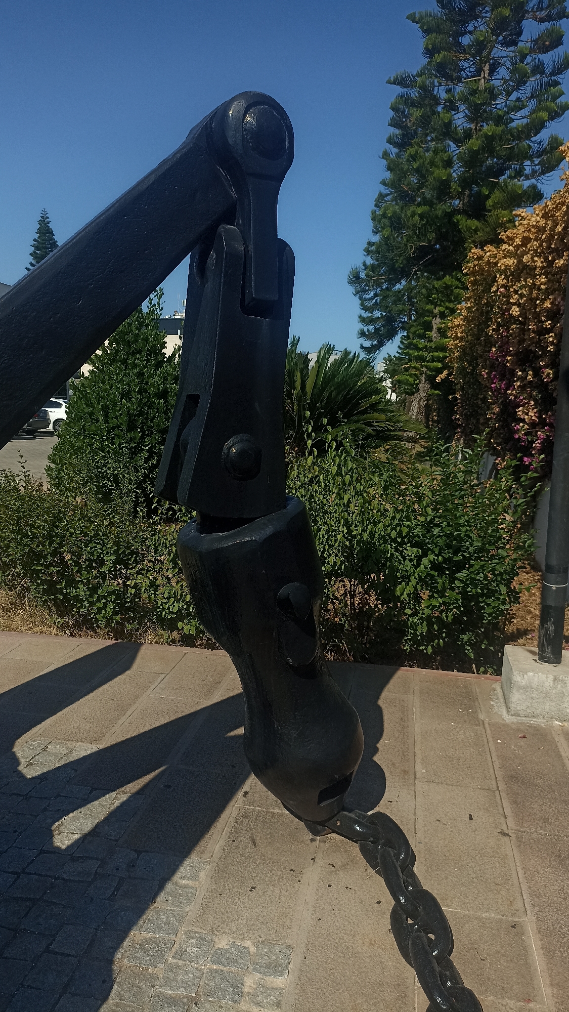 The image shows a close-up, angled view of a large, black metal anchor and a section of its chain. The anchor is positioned on a paved surface made of grey rectangular stones and smaller grey cobblestones. The anchor itself is dark, with visible joints and a large, curved fluke. A thick, black chain extends from the anchor's fluke and lies on the paving stones, casting long shadows. In the background, there are lush green bushes, a palm tree, and a tall evergreen tree, all set against a clear blue sky. To the left, a white car is partially visible behind the bushes, and a building can be seen in the distance. The lighting suggests a sunny day, with strong shadows cast by the anchor.